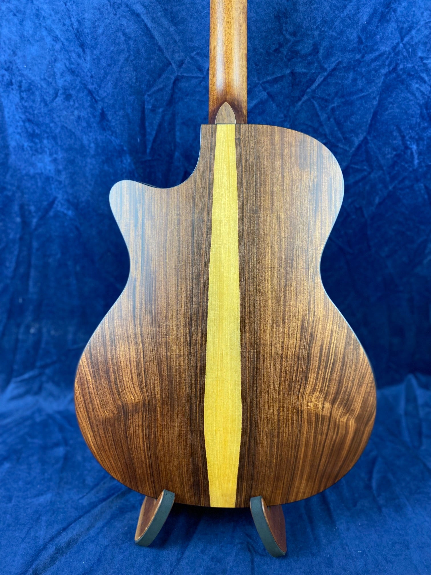 Acoustic guitar back with dark wood grain and a striking, wide vertical stripe of lighter wood running down the center, against a blue fabric background.