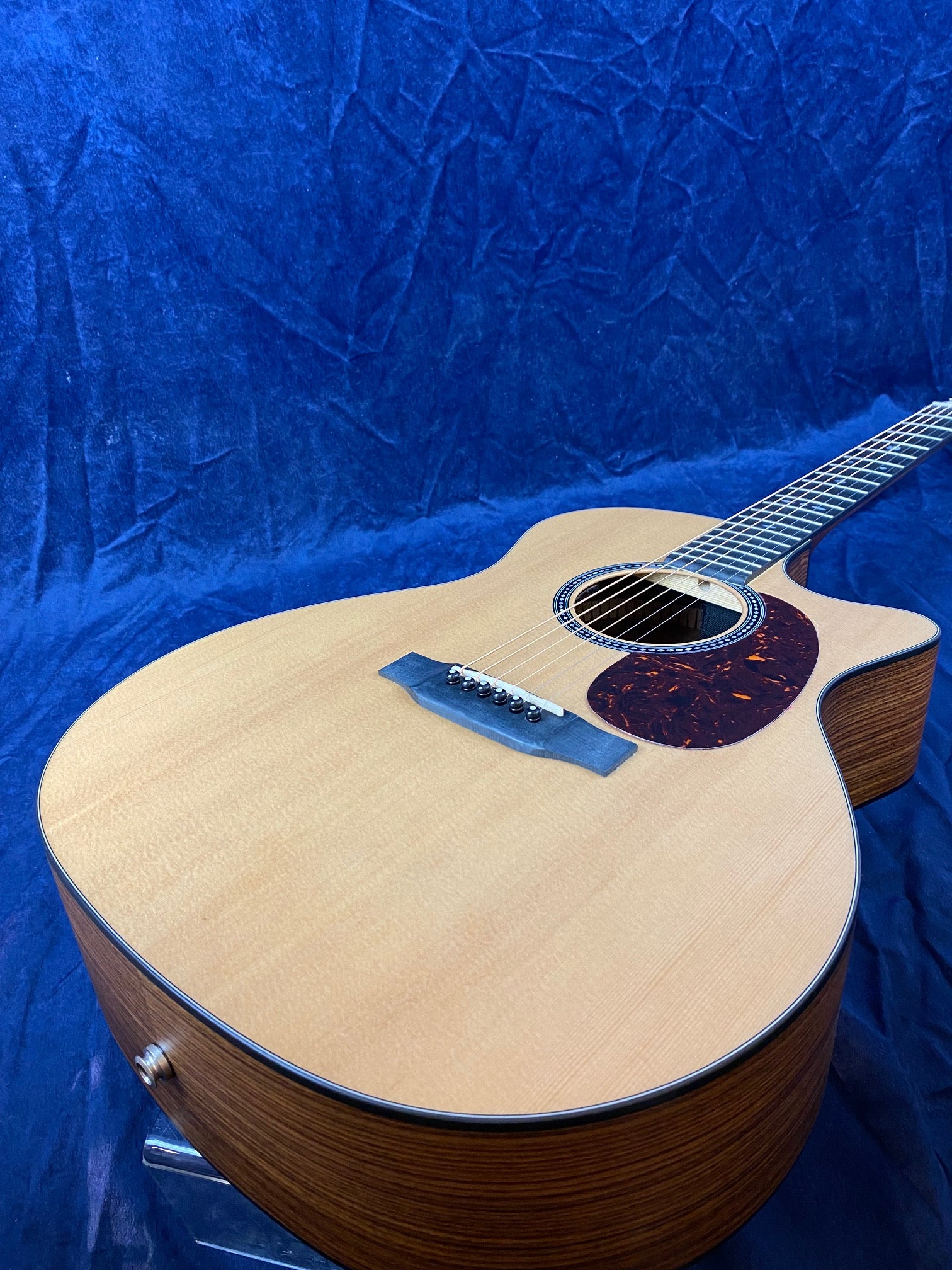Light wood acoustic guitar with a cutaway body, dark pickguard, six strings, and visible bridge, set against a deep blue textured background.