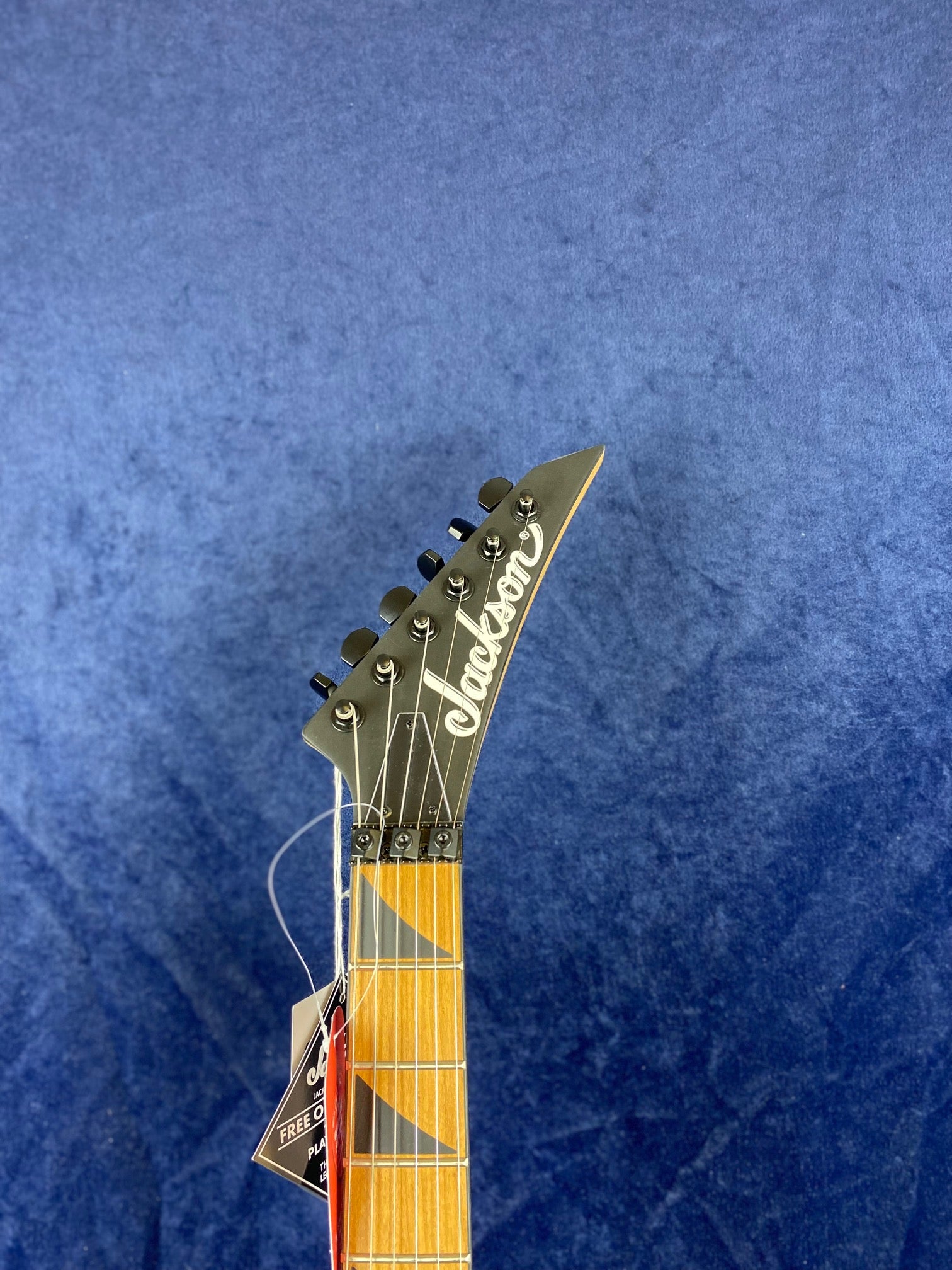 Jackson electric guitar headstock with six tuning pegs, maple neck, and tags attached, set against a blue background.