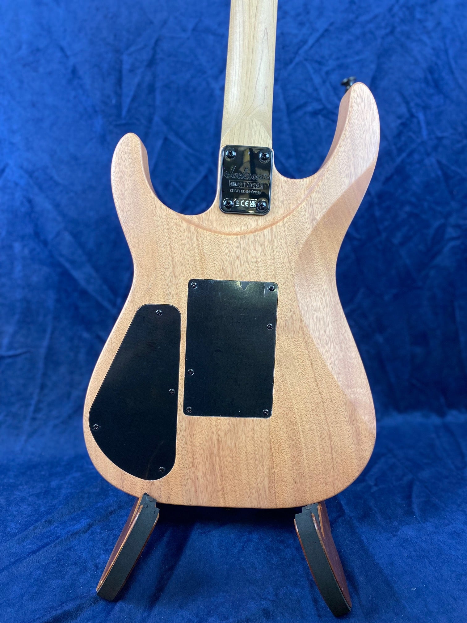 Light wood electric guitar body with two black plastic covers on the back, standing on a wooden stand against a blue background.