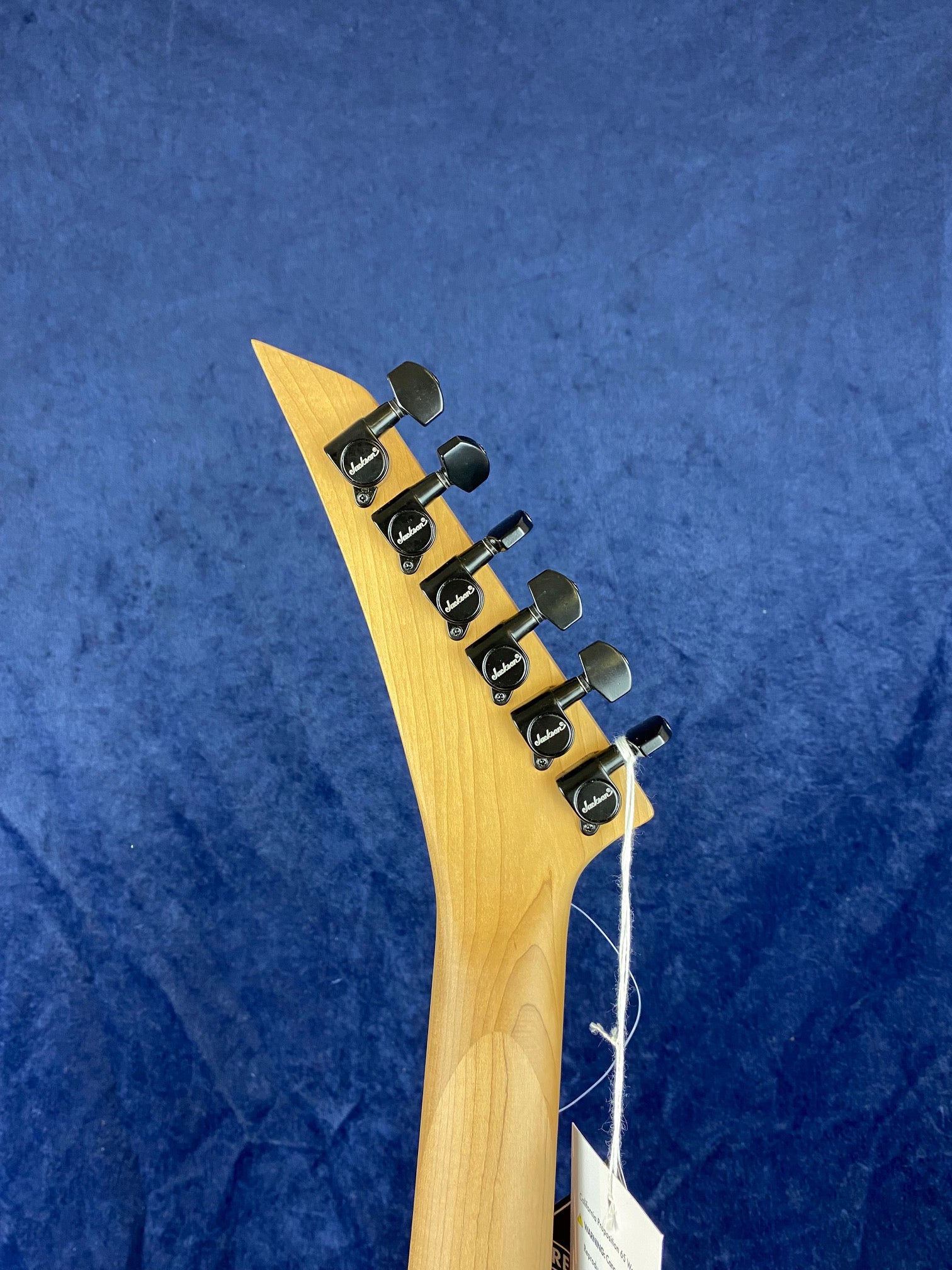 Natural wood-finish electric guitar headstock with six black Jackson tuners arranged in a line, set against a blue background.