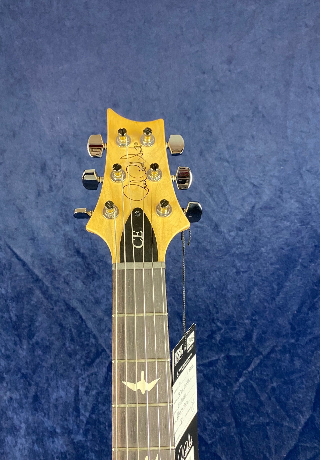 Six-string electric guitar headstock with chrome tuning pegs, signature, "CE" label, and a tag hanging from one tuner against a blue background.