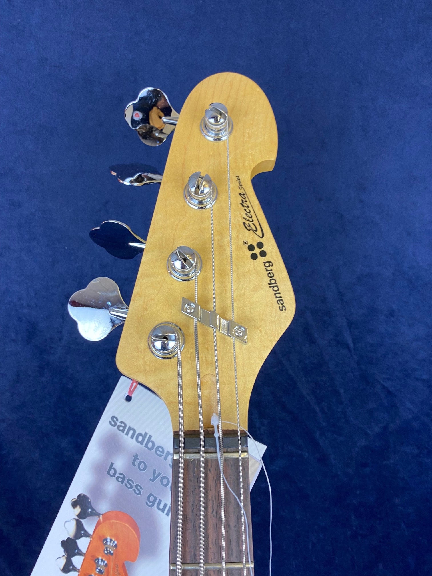 Headstock of a Sandberg Electra Series electric bass with four tuning pegs, metal string retainer, and attached promotional tag.