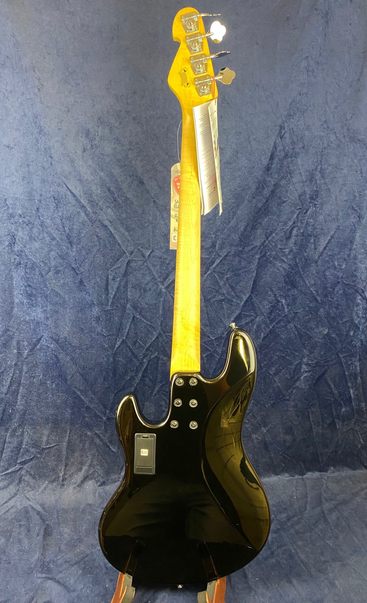 Electric bass shown from the back; glossy black body, light wood neck, four tuning pegs, and a battery compartment cover on the body.