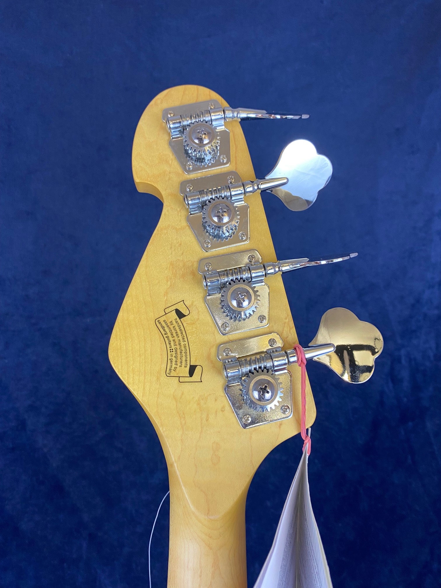 Four metal tuning pegs and gear mechanisms on a natural wood electric bass headstock, with a tag attached by a pink rubber band.