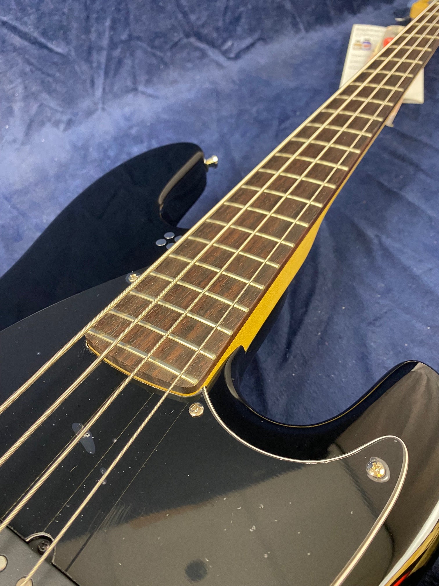 Close-up of a black electric bass guitar showing the strings, frets, and part of the dark wooden fingerboard against a blue textured background.