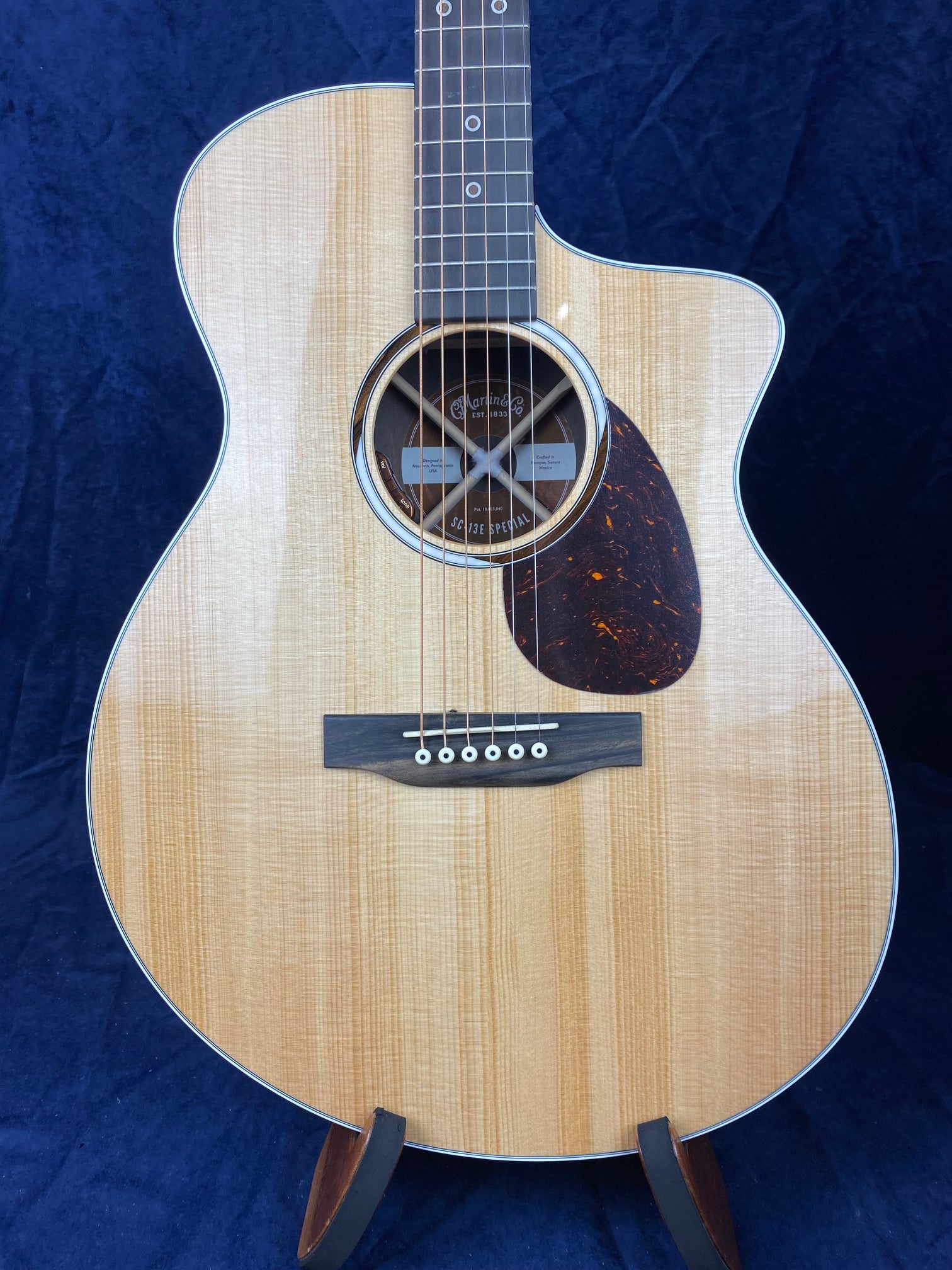 Light wood acoustic guitar with a cutaway body, six strings, dark pickguard, and circular soundhole, displayed upright on a stand against dark fabric.