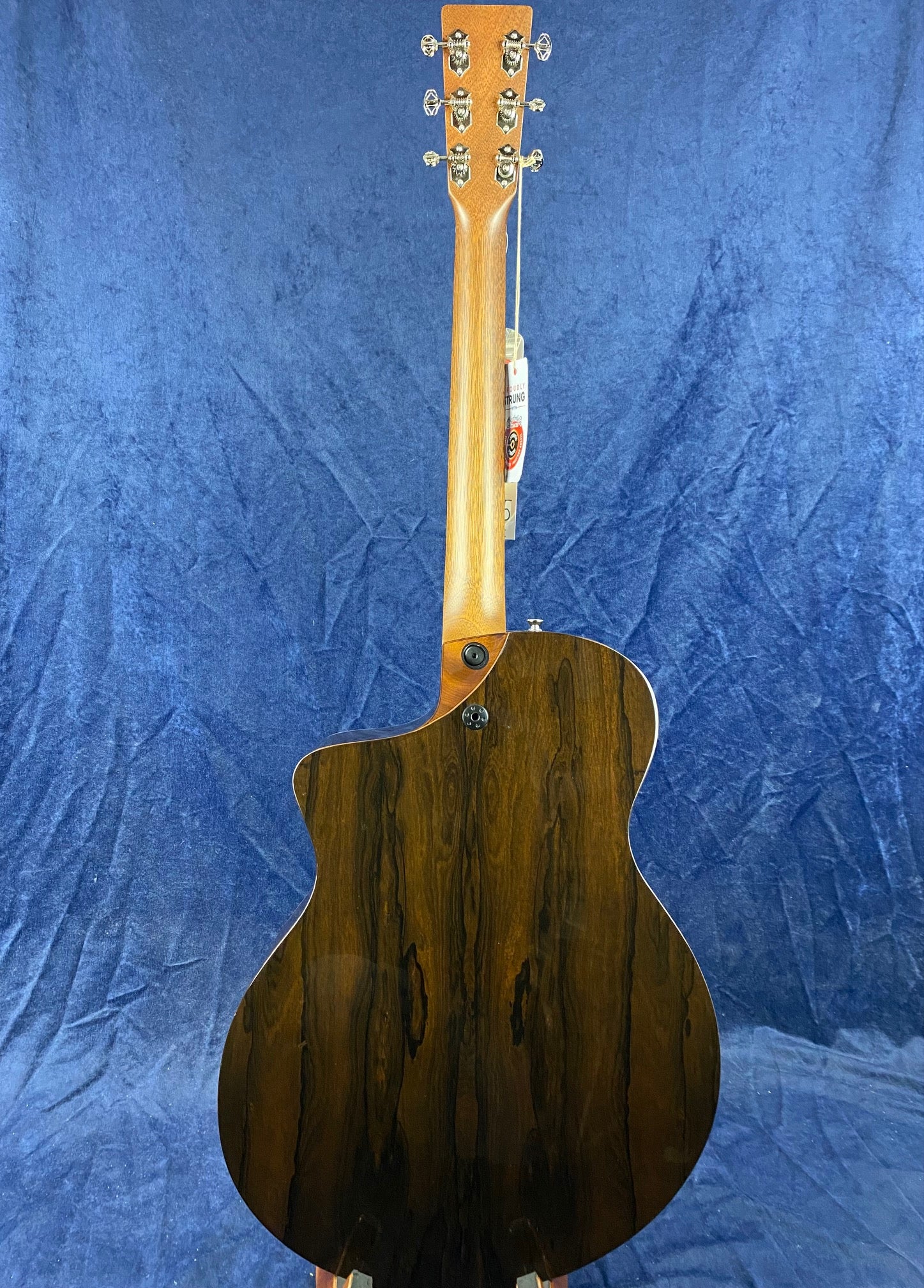 Acoustic guitar shown from the back, featuring a glossy dark wood body with natural grain patterns and a lighter wood neck against a blue background.