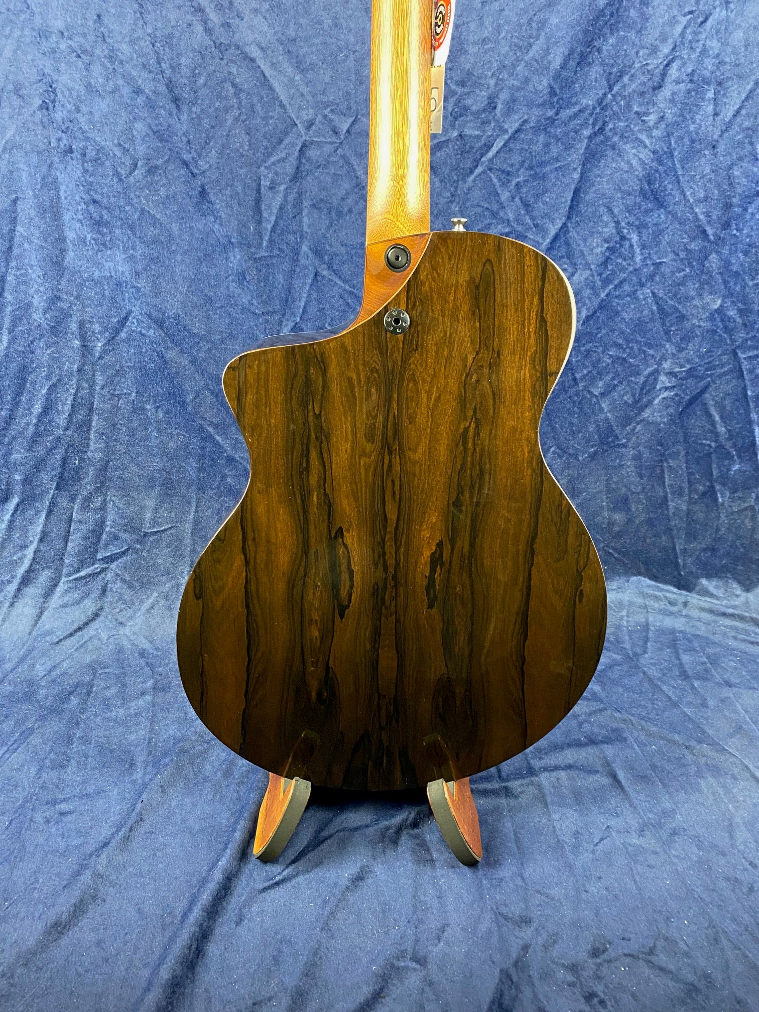 The back of an acoustic guitar with dark wood grain patterns and a light-colored neck, standing upright on a blue textured cloth background.