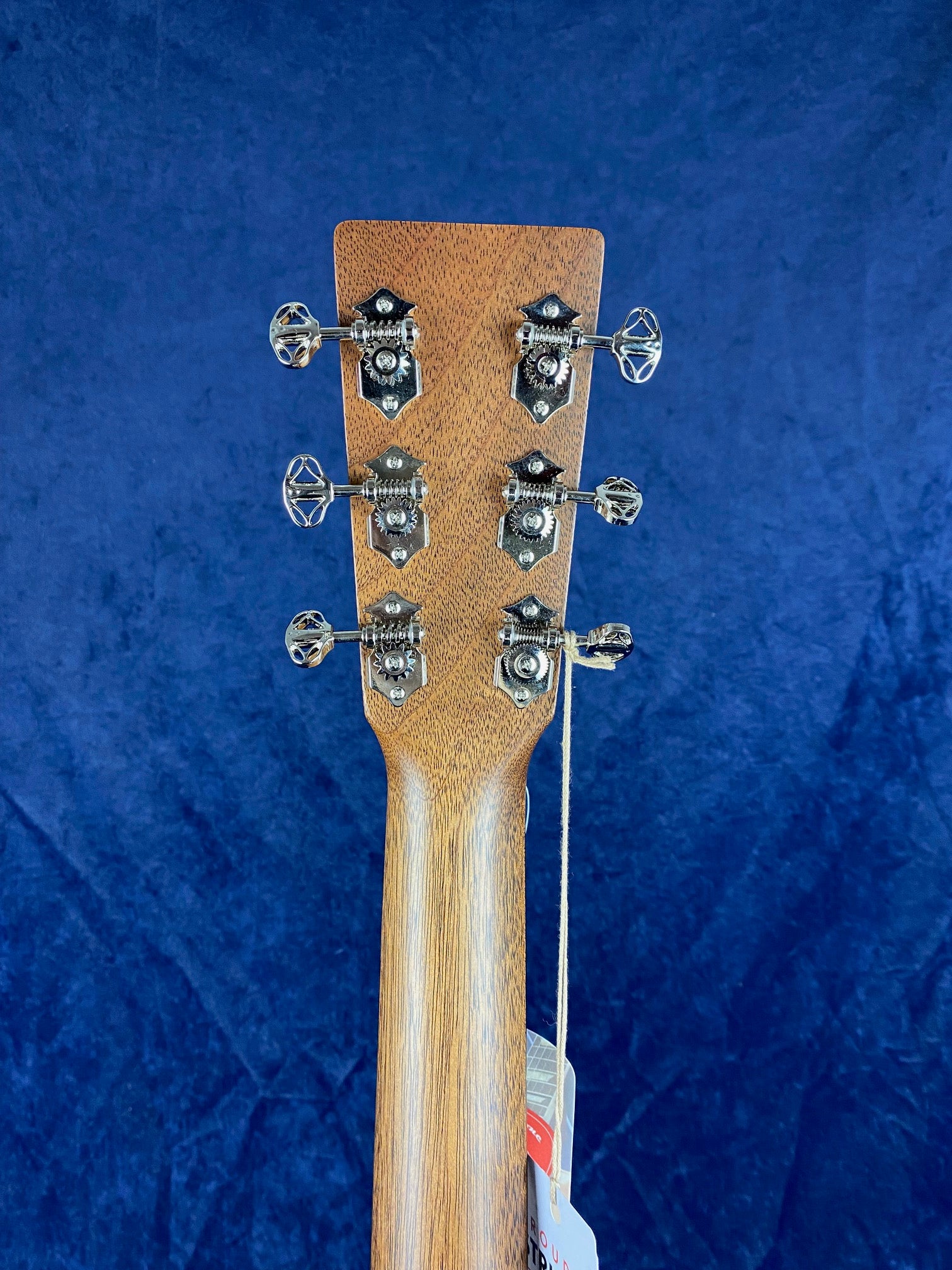 Back of an acoustic guitar headstock showing six metal tuning pegs and a wooden neck, with product tags hanging from one tuning peg.