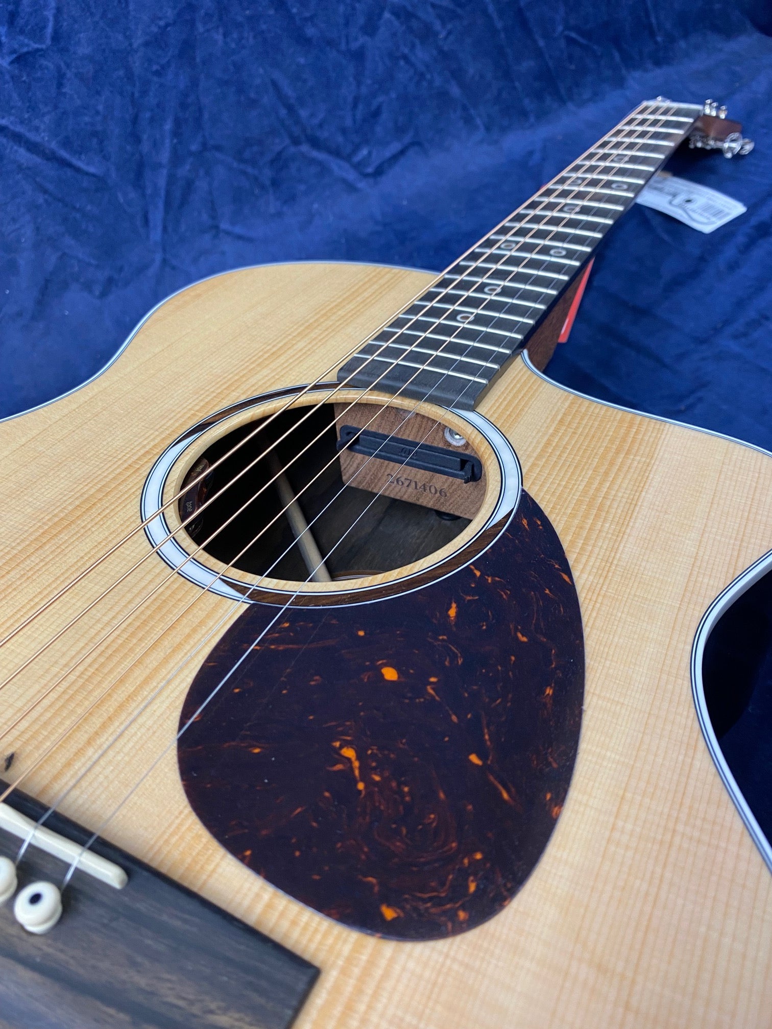 Light wood acoustic guitar with a tortoiseshell-style pickguard, six steel strings, and a visible soundhole pickup under a blue fabric background.