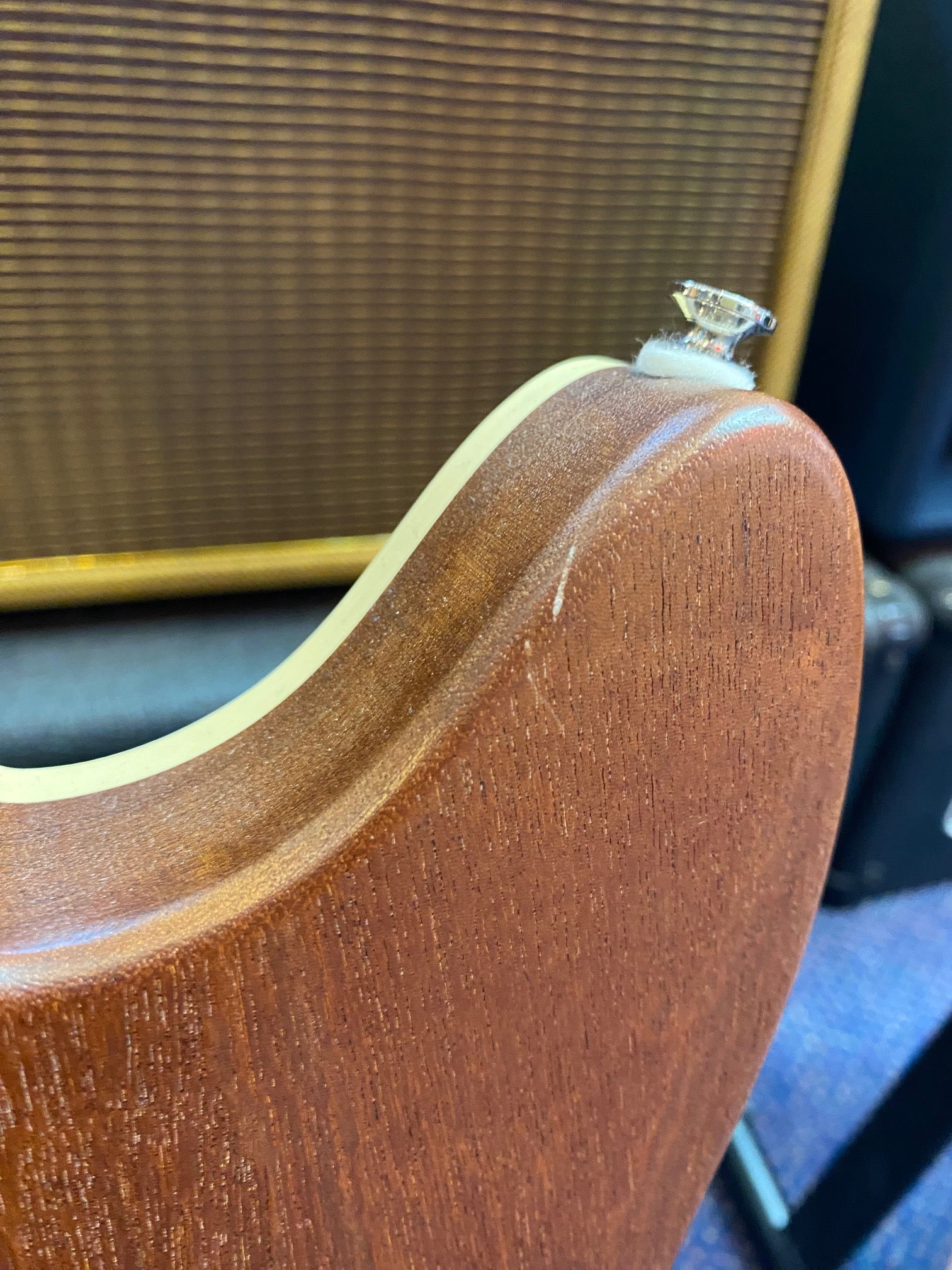 Upper edge of a brown electric guitar body with cream binding, showing a strap button and a small surface scratch. Background includes an amplifier grille.