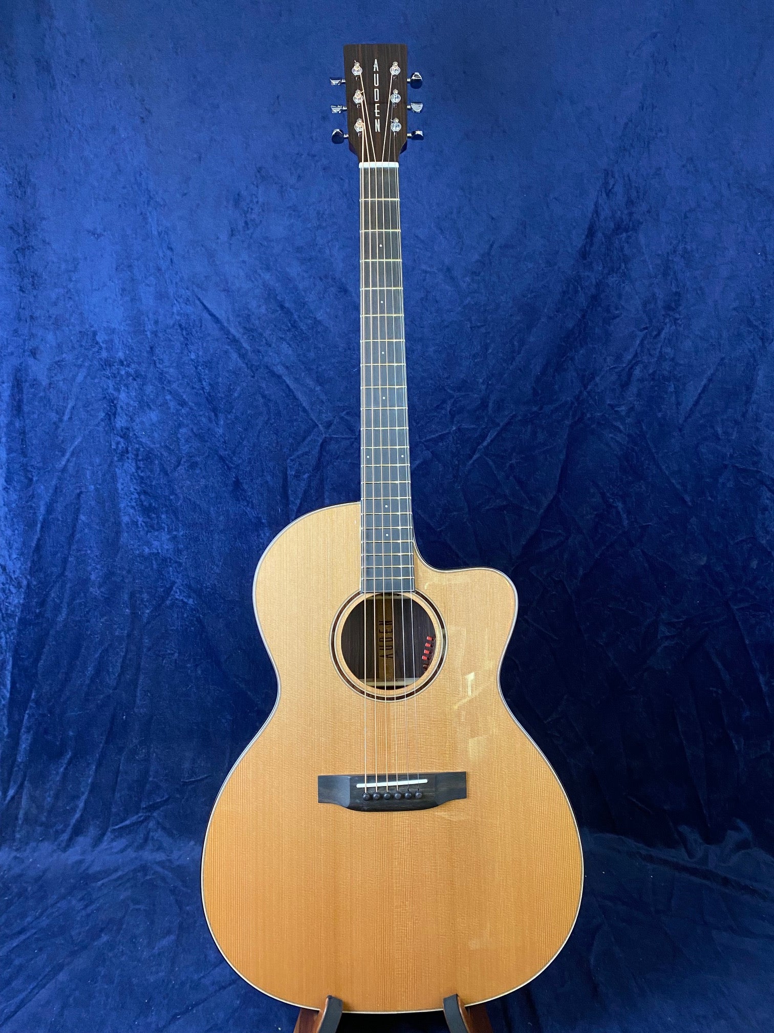 Natural wood acoustic guitar with a cutaway body shape, six strings, and “Auden” logo on the headstock, set against a blue background.