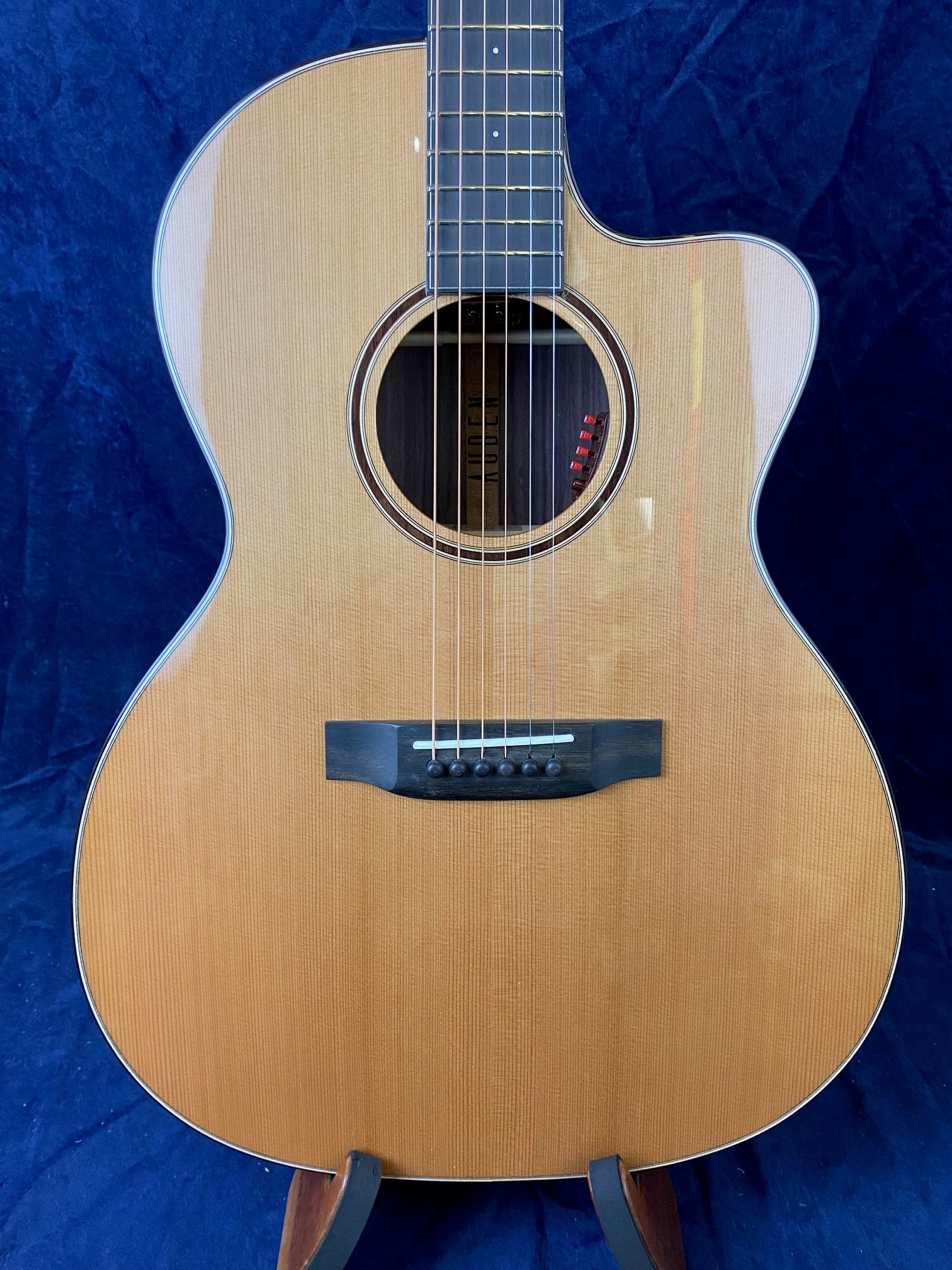 Glossy light wood acoustic guitar with a cutaway body shape, six strings, and black bridge, positioned upright against a dark blue background.