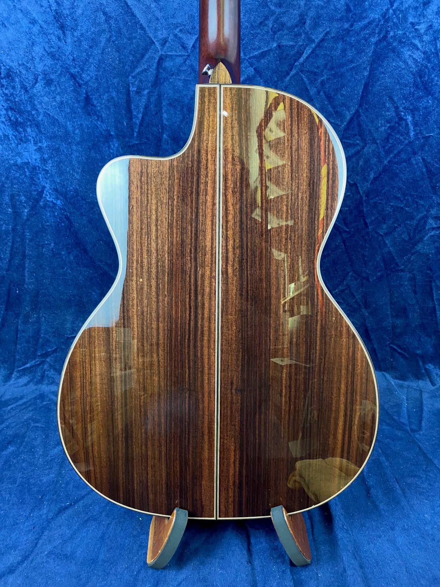 A high-gloss acoustic guitar back with dark brown wood grain, divided by a narrow light strip down the center, resting on a stand.