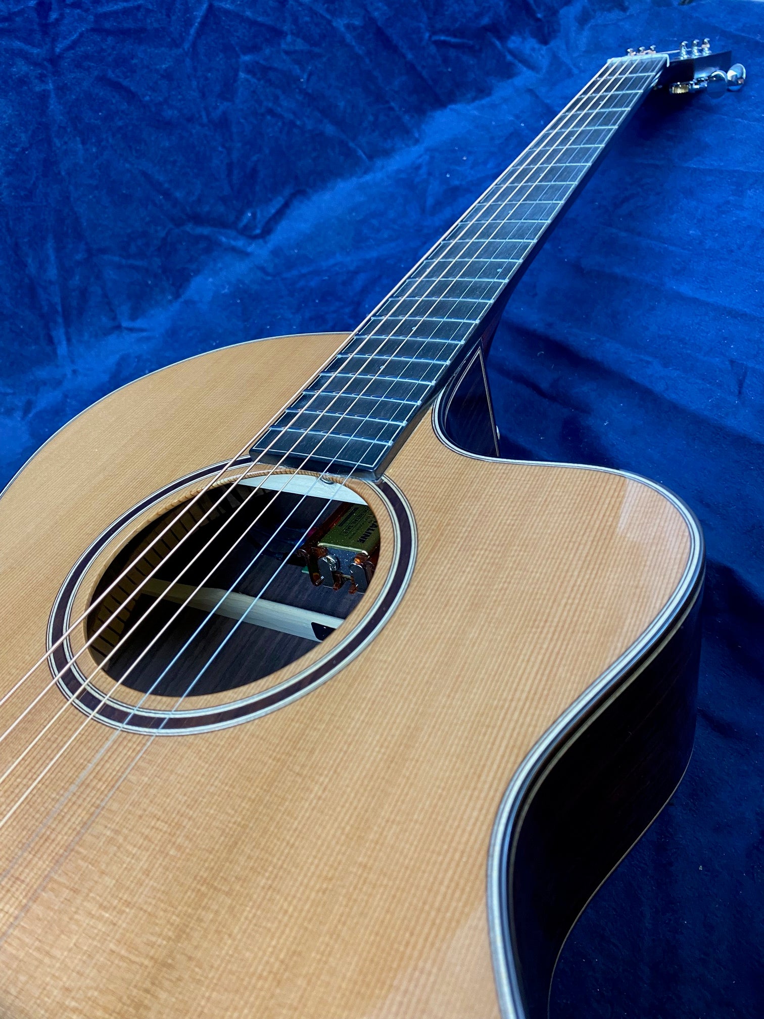 Light wood acoustic guitar with a round sound hole, dark fretboard, and metal tuning pegs, positioned on a deep blue fabric background.