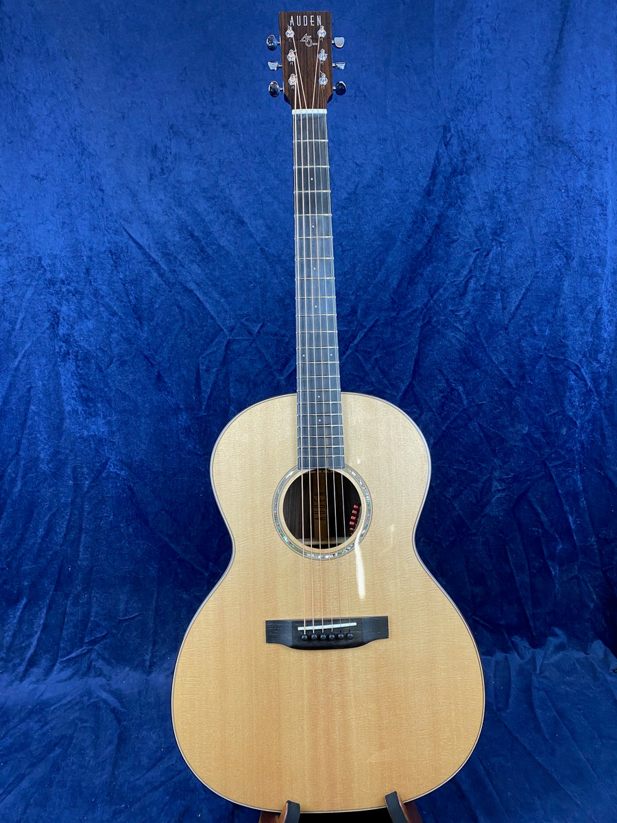 Light wood Auden acoustic guitar with six strings, dark wood fretboard, and round soundhole, displayed upright against a blue textured background.