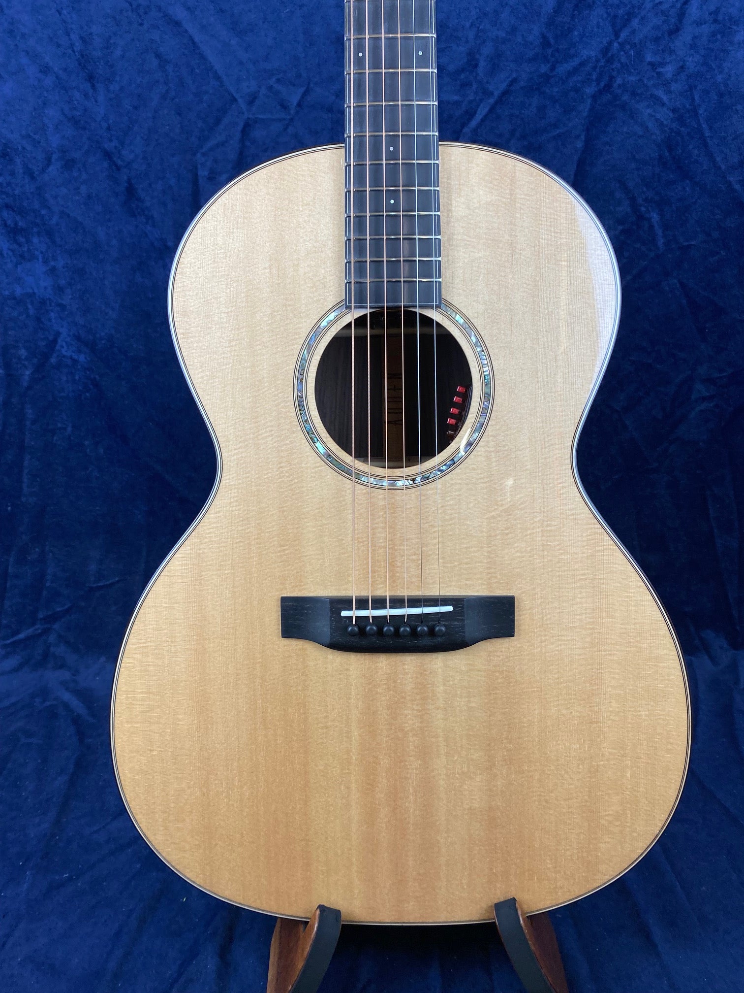 Light wood acoustic guitar with a rounded body, abalone rosette around the soundhole, black bridge, and six strings against a dark blue background.