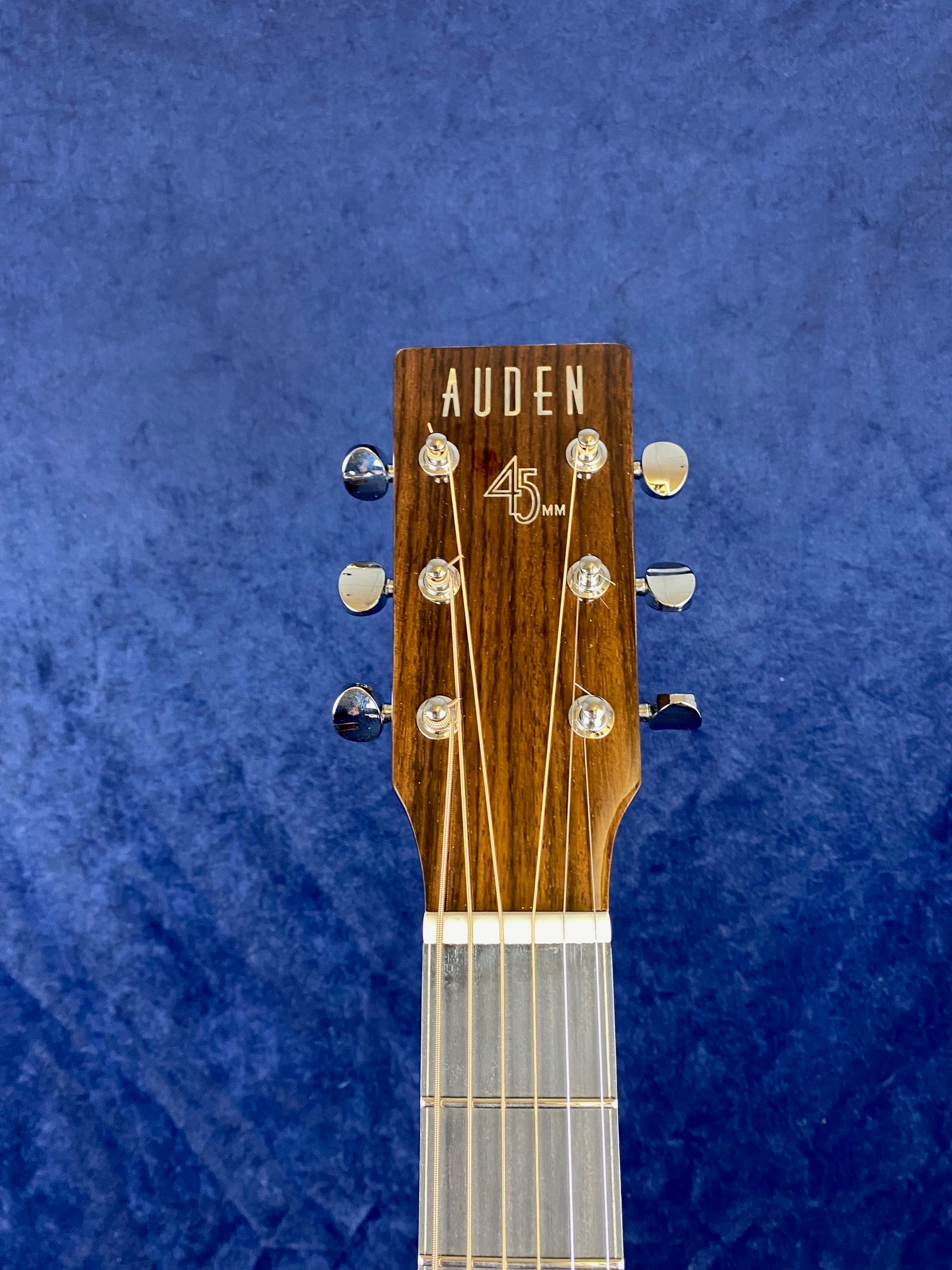 Six-string acoustic guitar headstock with chrome tuning pegs, "AUDEN" logo, and "45MM" printed on dark wood, set against a blue background.