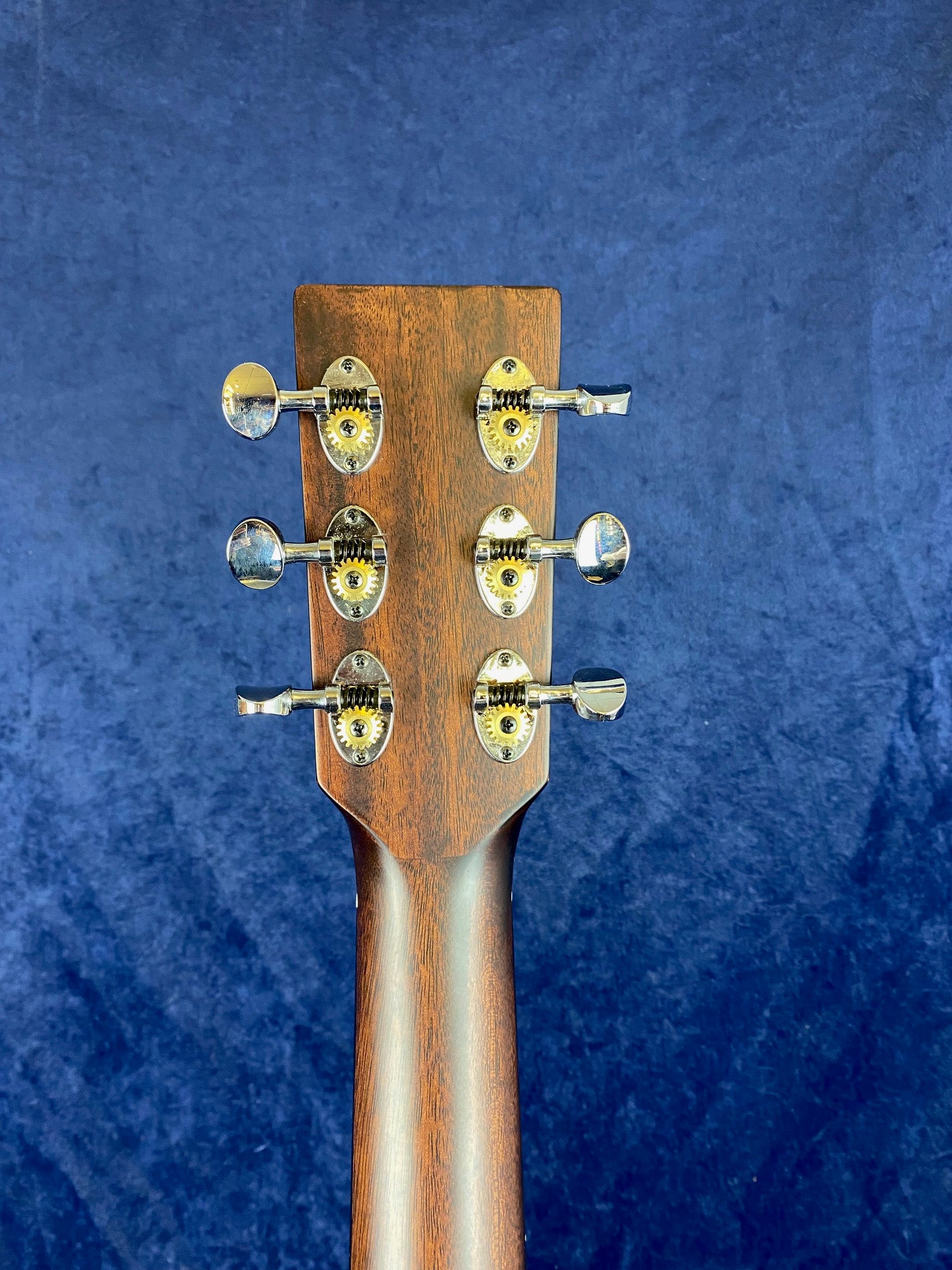 Back of an acoustic guitar headstock showing six chrome tuning pegs and a wooden neck against a dark blue background.