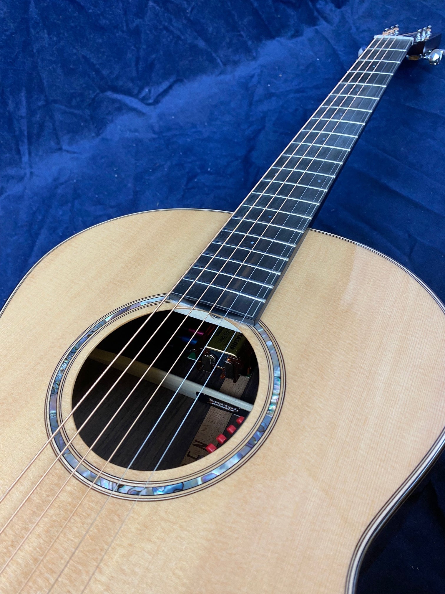Light wood acoustic guitar with a round sound hole, abalone inlay around the hole, and six steel strings resting on a dark blue fabric background.