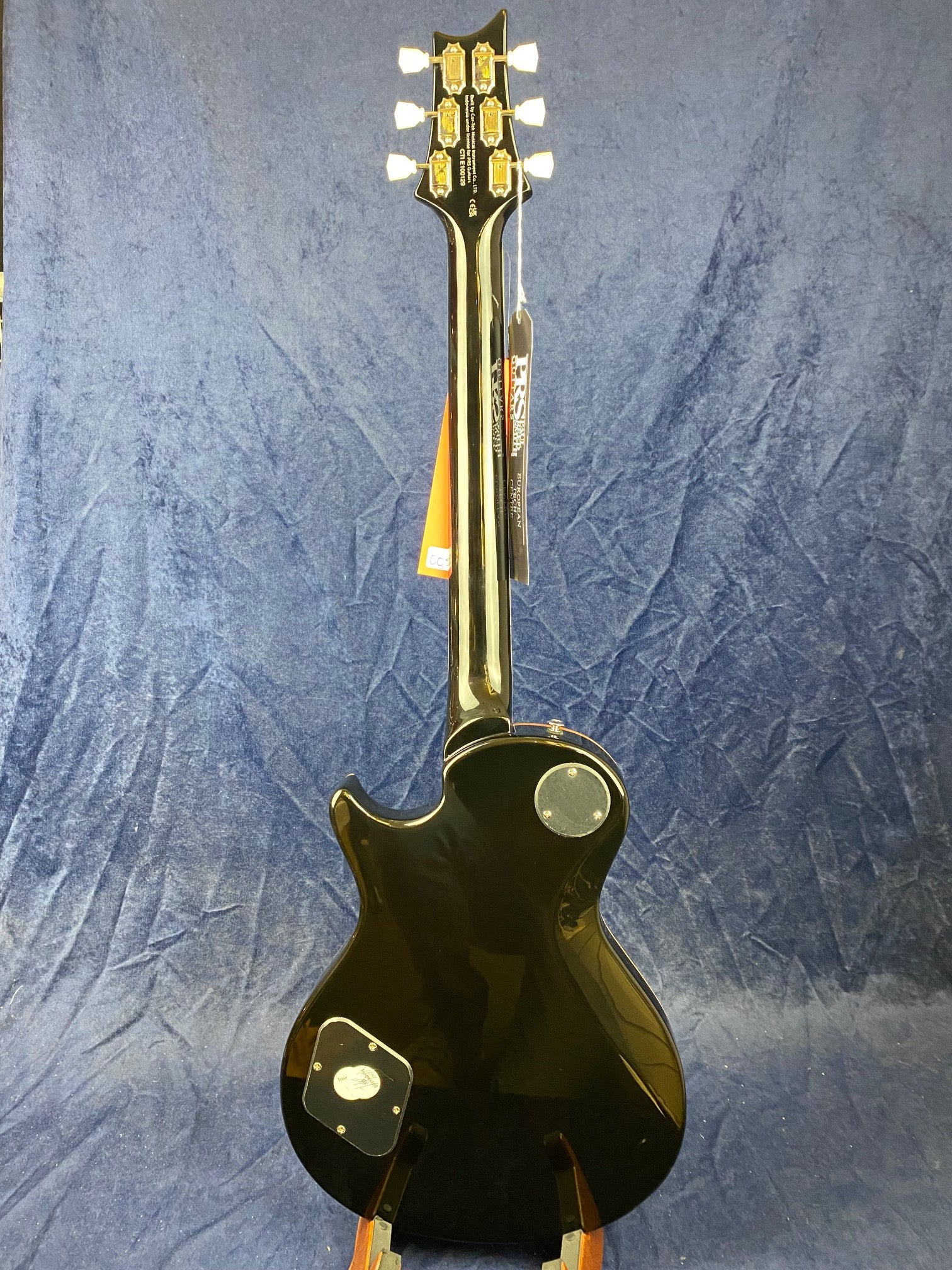 Glossy black electric guitar shown from the back, with white tuning pegs, two round covers on the body, and hang tags on the neck.