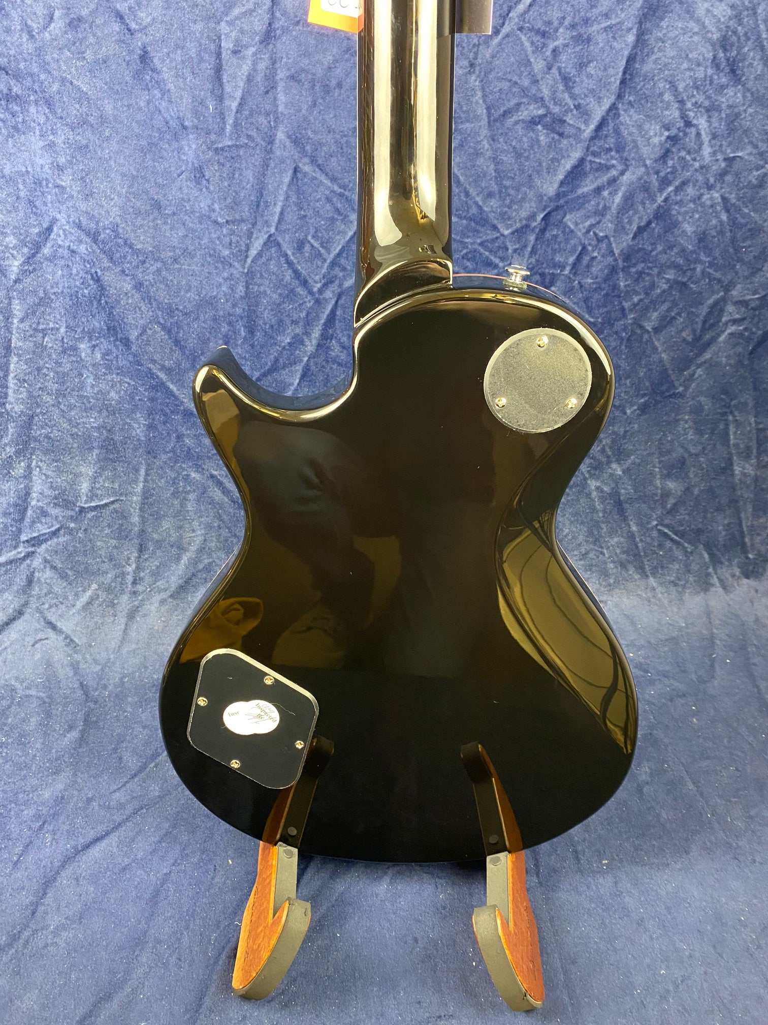 Glossy black electric guitar shown from the back, with round and square control cavity covers and a single cutaway body shape.
