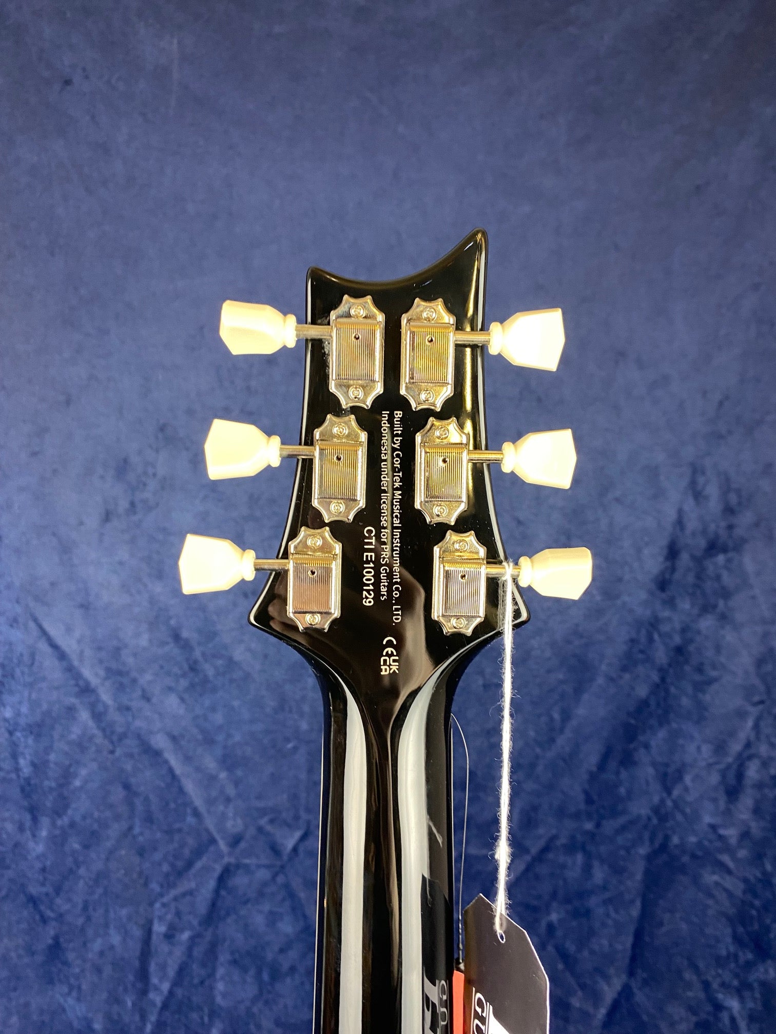 Back of electric guitar headstock with six vintage-style tuning pegs and product tags hanging from a white string against a blue background.