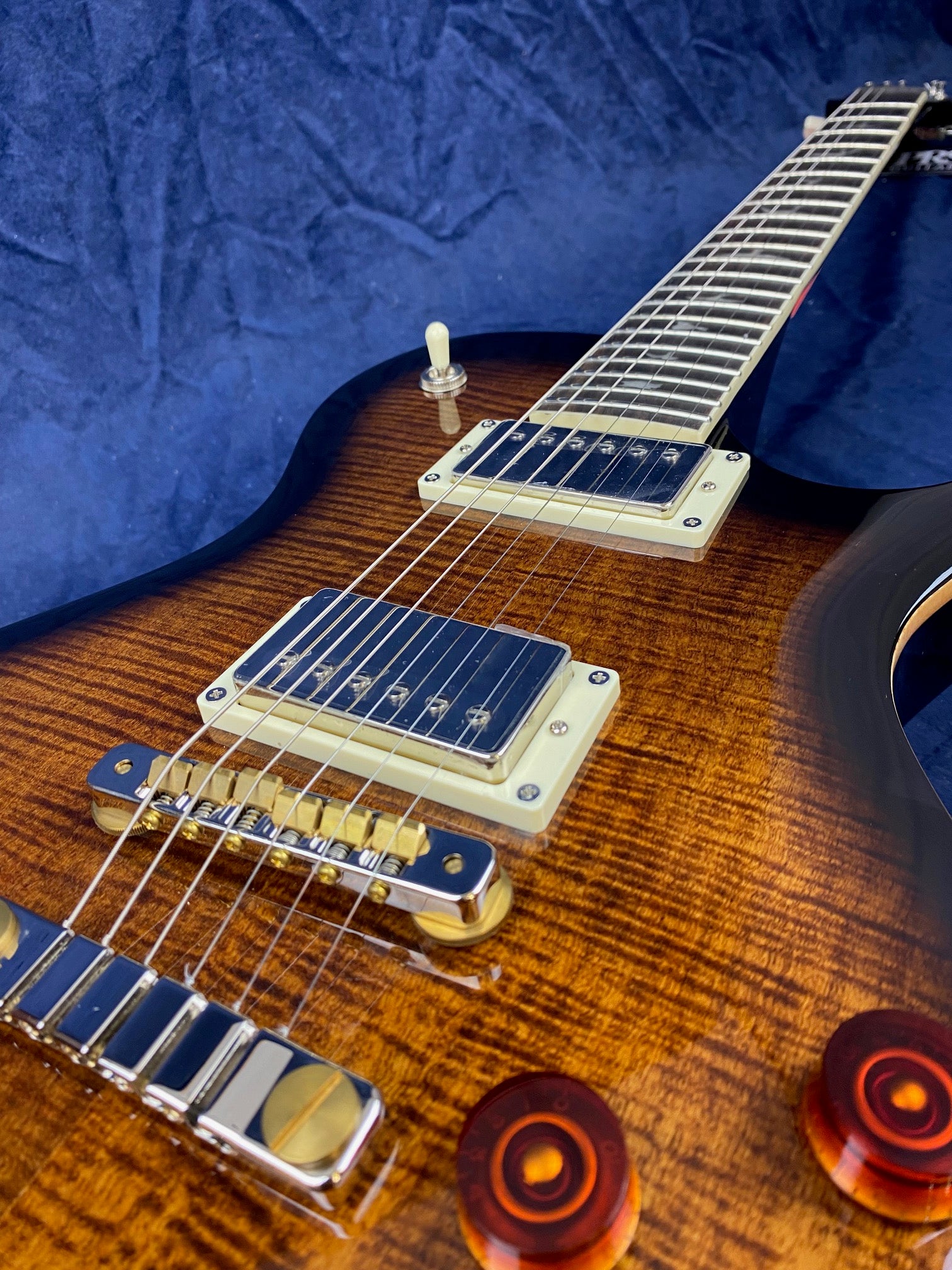Sunburst electric guitar with a glossy flame maple top, gold hardware, dual humbucker pickups, and amber control knobs against a dark blue background.
