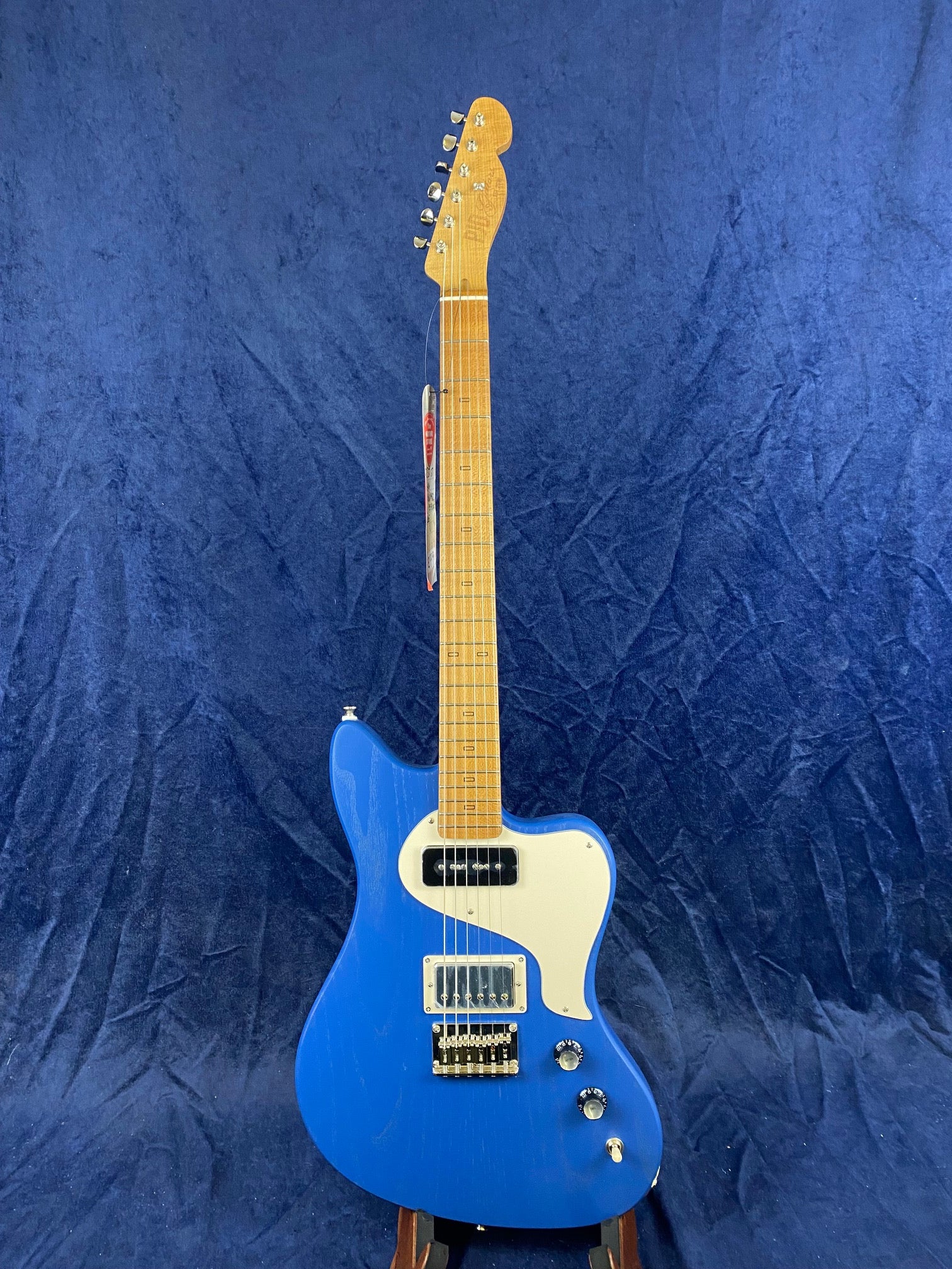 Electric guitar with a blue body, maple neck, two pickups, a white pickguard, three control knobs, and a switch, displayed against a blue background.