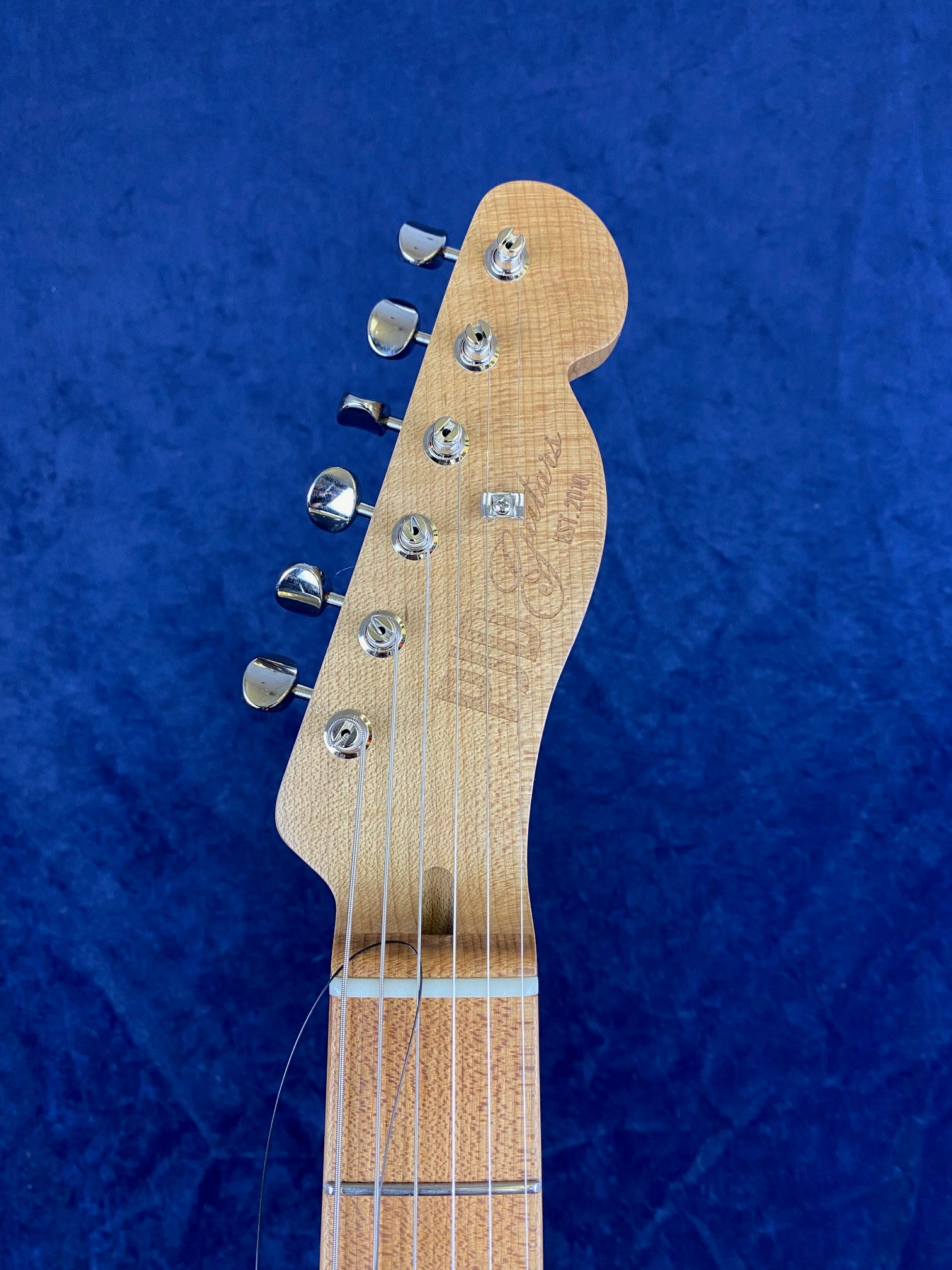 Light wood electric guitar headstock with six chrome tuning pegs, visible strings, and faint cursive text, against a deep blue background.