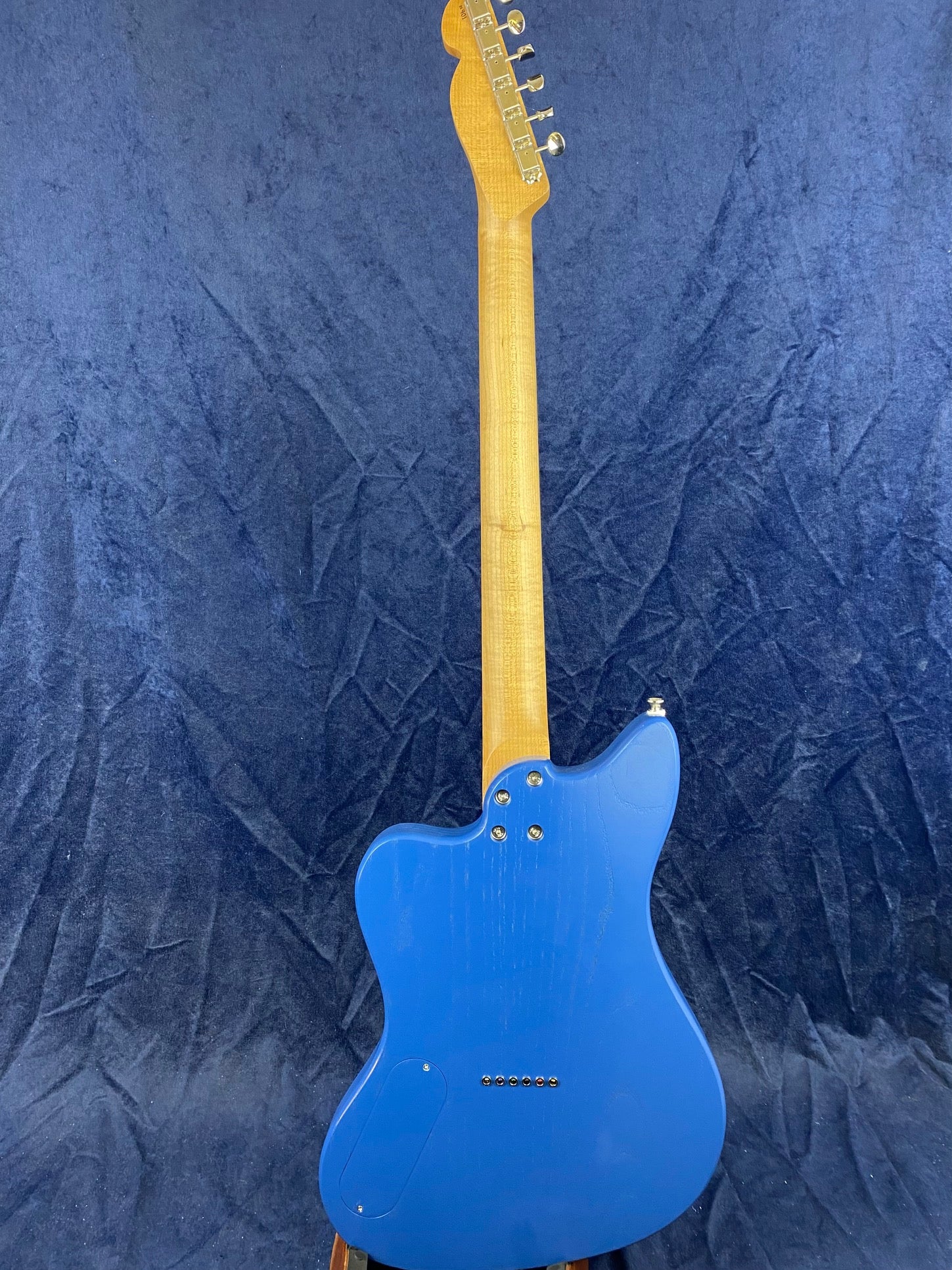 Electric guitar shown from the back, featuring a bright blue body and a natural wood neck with six tuning pegs on the headstock.