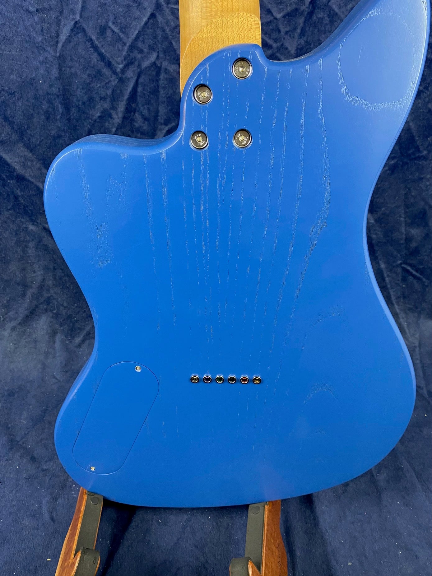 Back of a blue electric guitar body with visible wood grain, four neck bolts, a rectangular electronics cover, and six ferrules for string-through design.