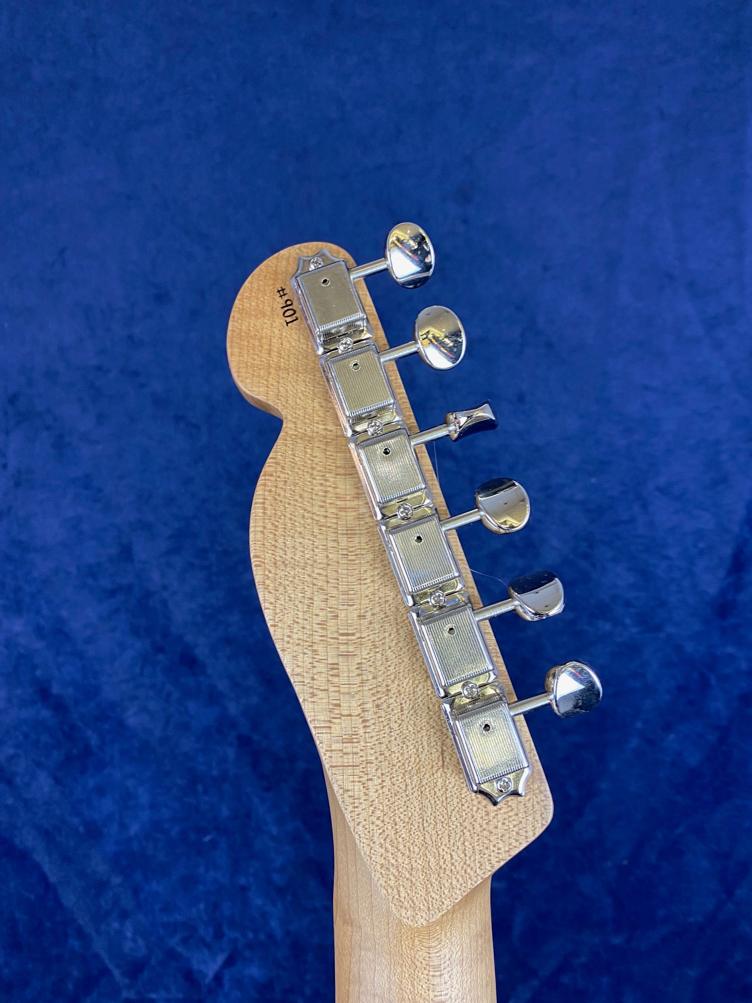 Close-up of an electric guitar headstock with six chrome tuning pegs on a light wood grain background, set against a blue backdrop.