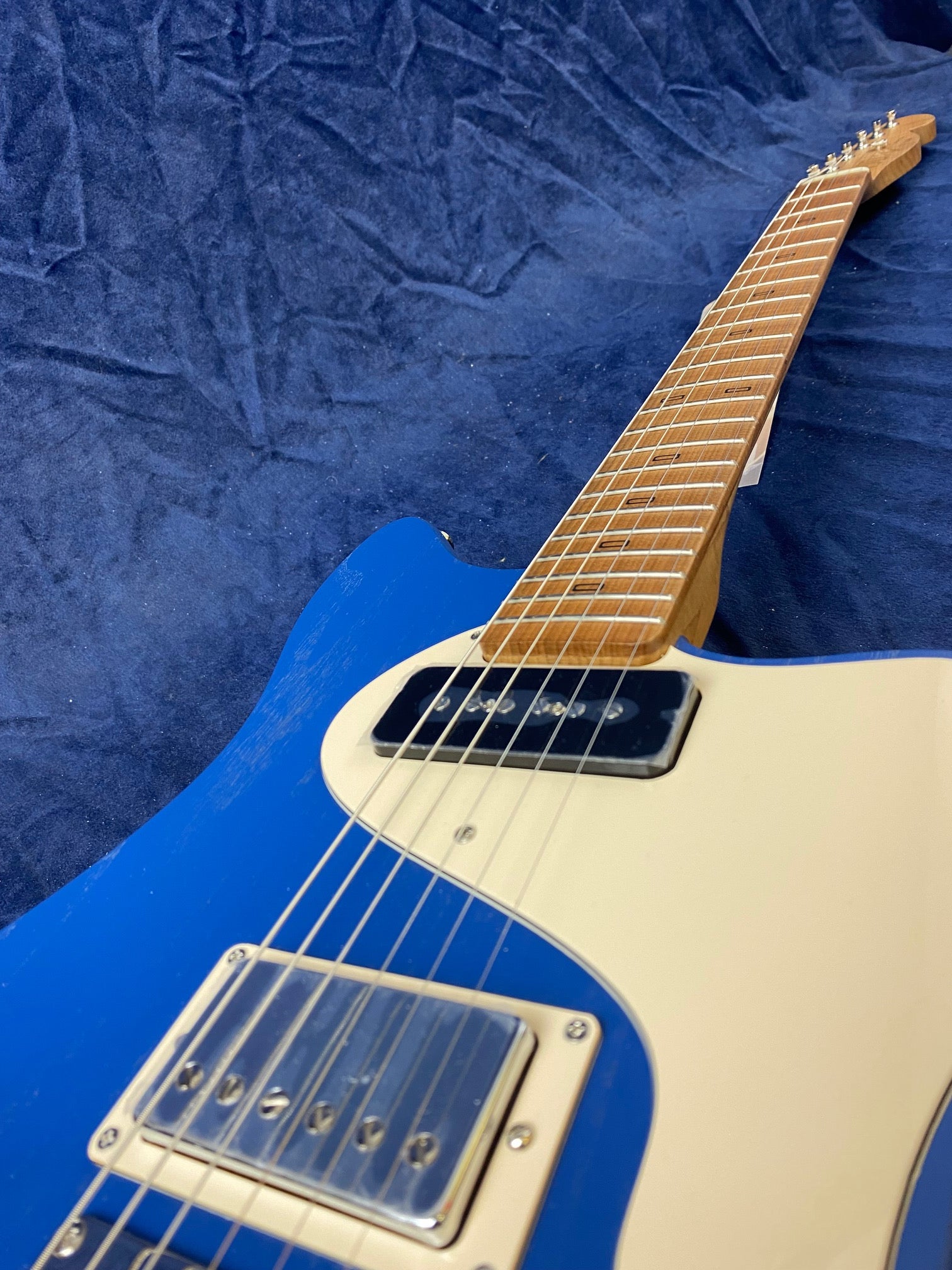 Electric guitar with a blue body, cream pickguard, chrome pickups, and a light wood neck, lying on a textured dark blue fabric.