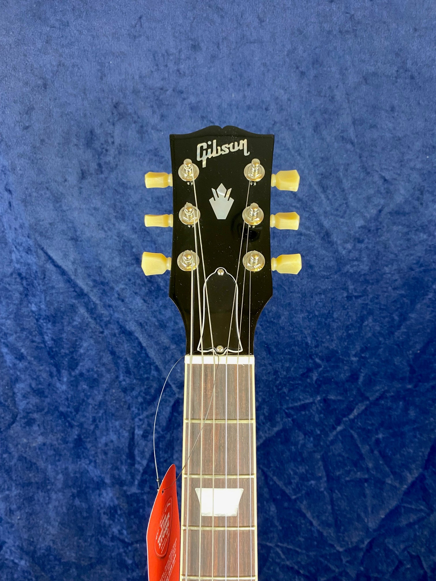 Black Gibson electric guitar headstock with six cream tuning pegs, mother-of-pearl inlay, and part of the fretboard visible against a blue background.