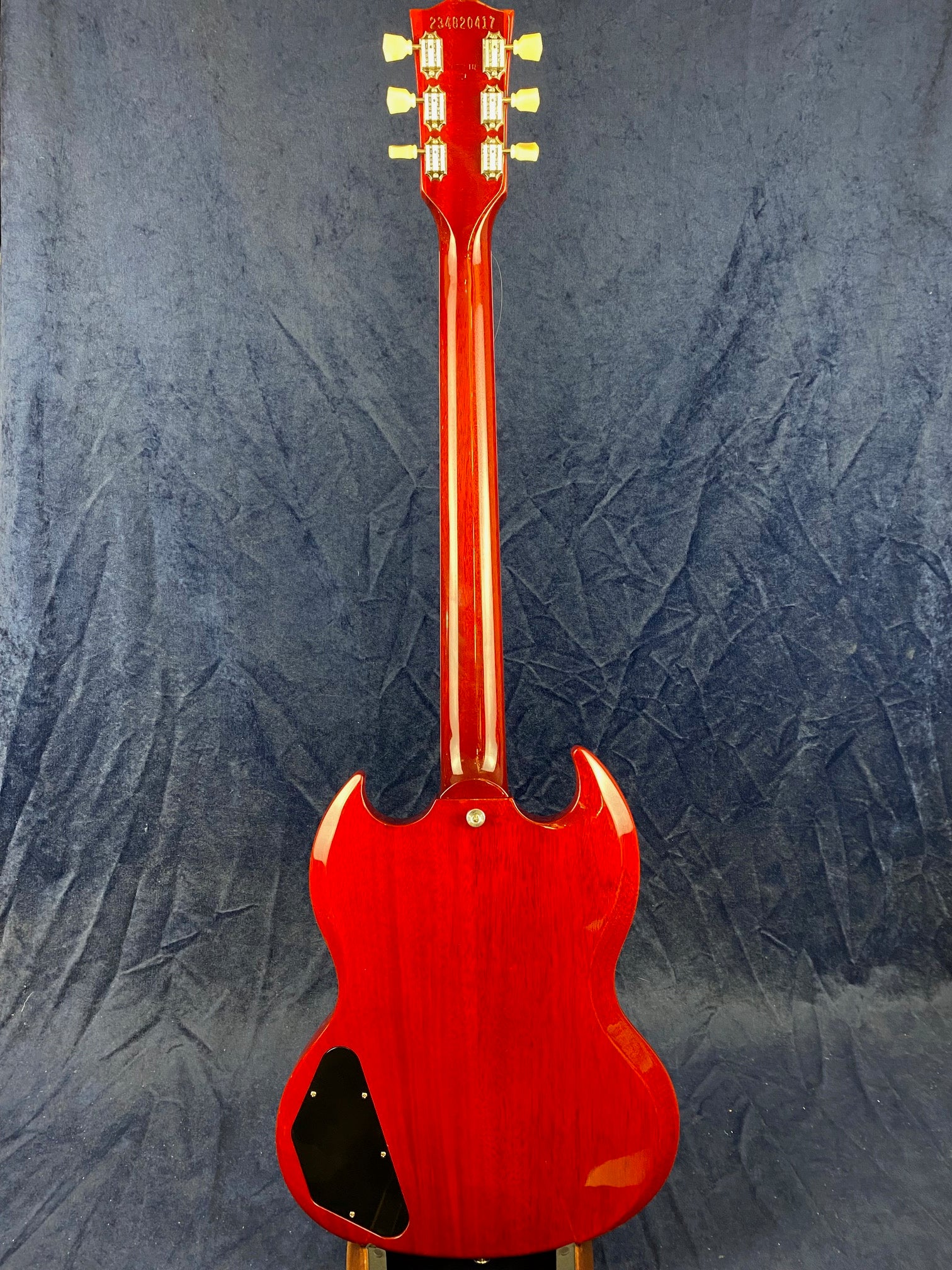 Back view of a red electric guitar with a double-cutaway body, six tuning pegs, and a black plastic cover on the lower left side of the body.