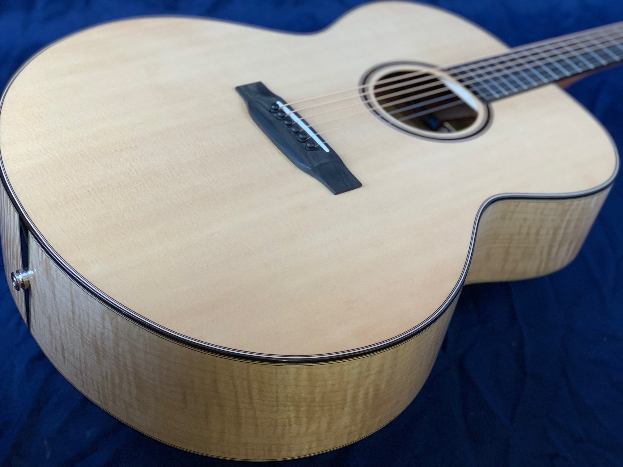 Light wood acoustic guitar with a glossy finish, dark bridge and rosette, and subtle flame pattern on the sides, resting on a dark blue surface.