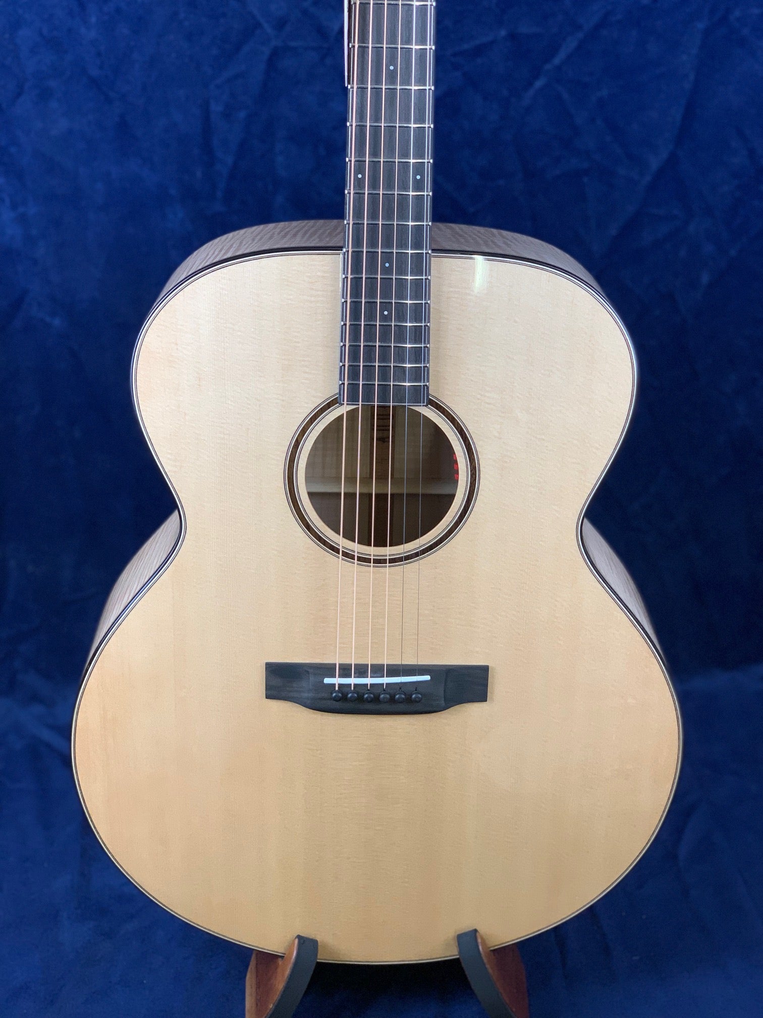 Light wood acoustic guitar with a round body, six strings, black bridge, and round sound hole, shown upright against a dark blue background.