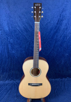 Auden acoustic guitar with a light wood top, dark sides, six strings, and a red tag hanging from the headstock against a blue background.