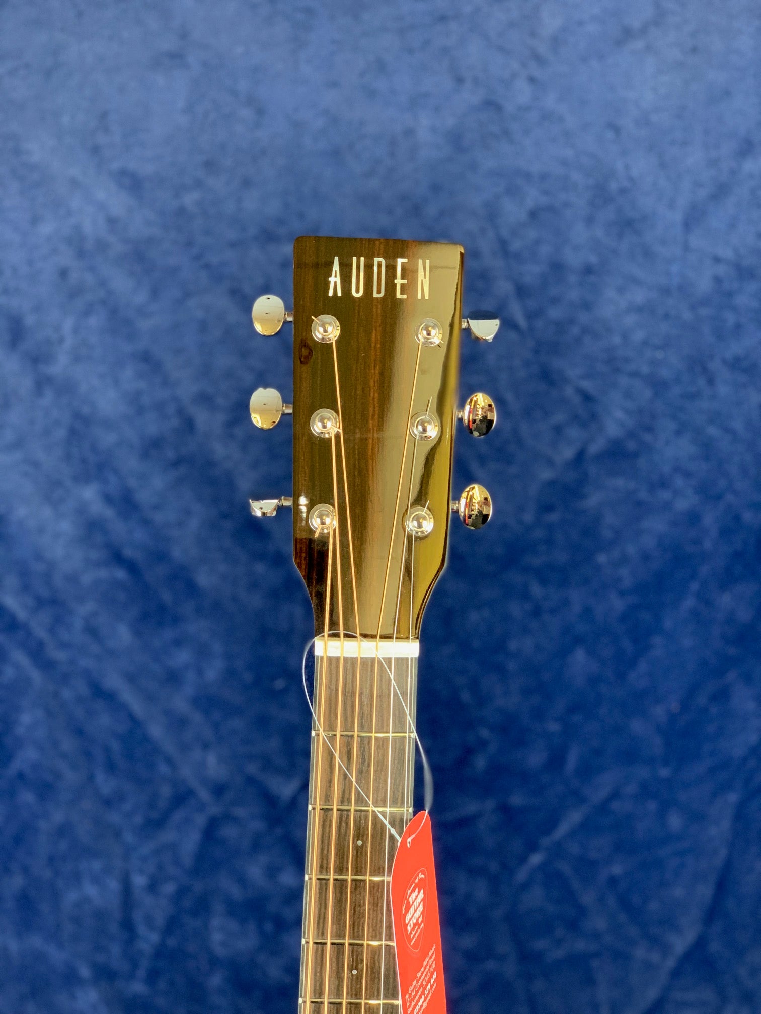 Auden acoustic guitar headstock with six tuning pegs, wood grain finish, and a red tag hanging from the neck against a blue background.
