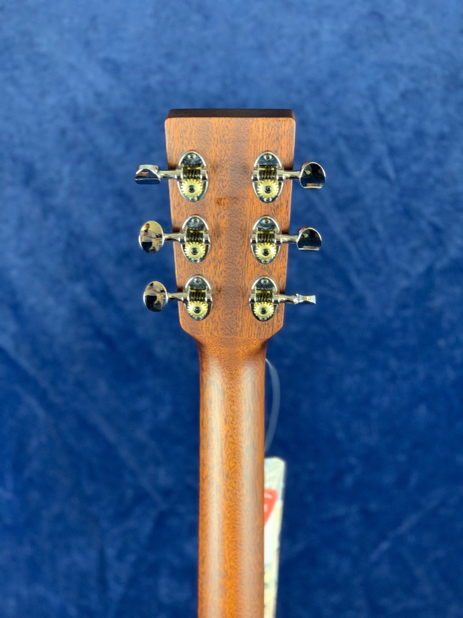 Back of an acoustic guitar headstock showing six chrome tuning pegs on a wooden neck, against a blue background.