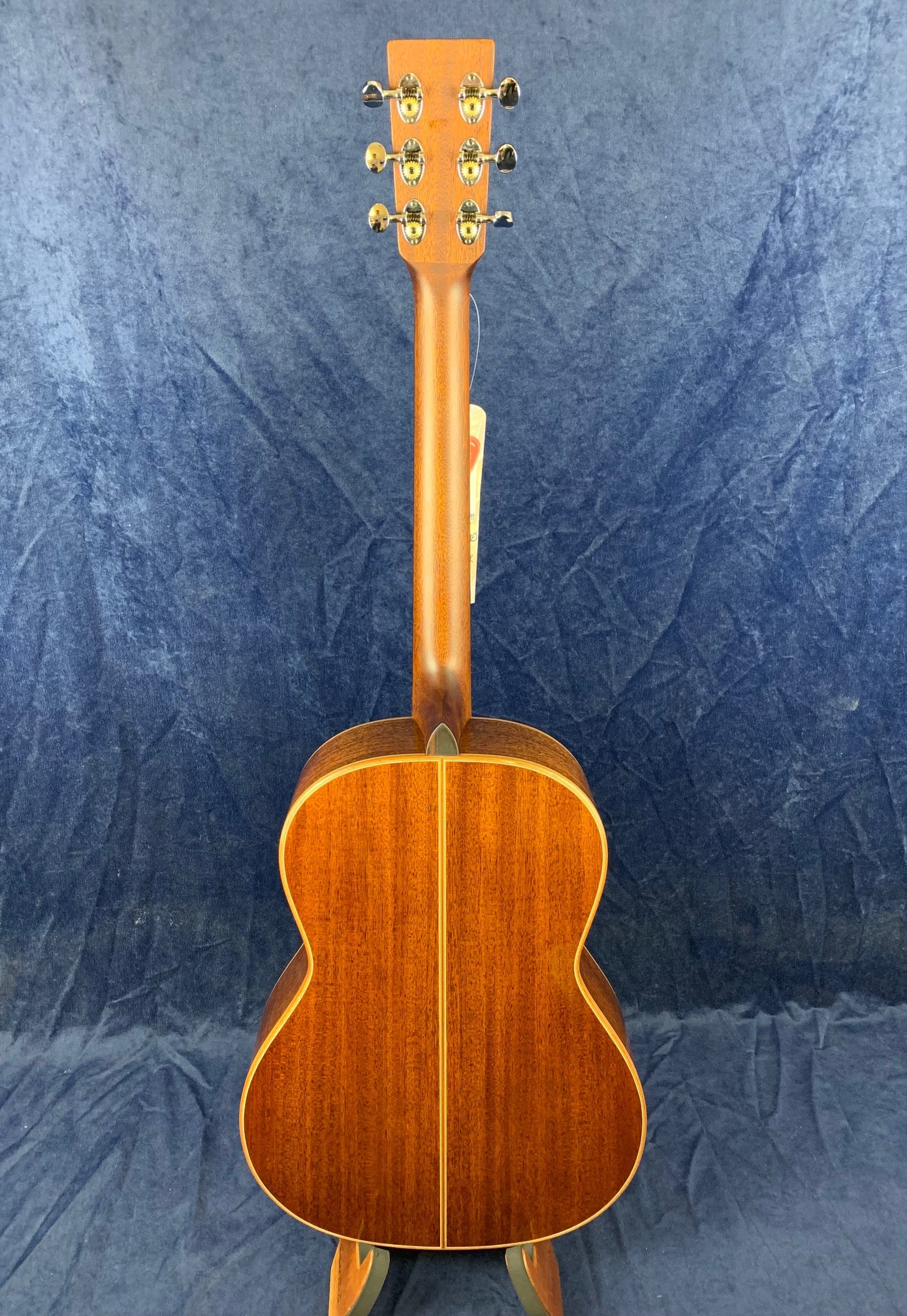 Acoustic guitar shown from the back, featuring a glossy, dark wood finish with a vertical center seam and gold tuning pegs on the headstock.
