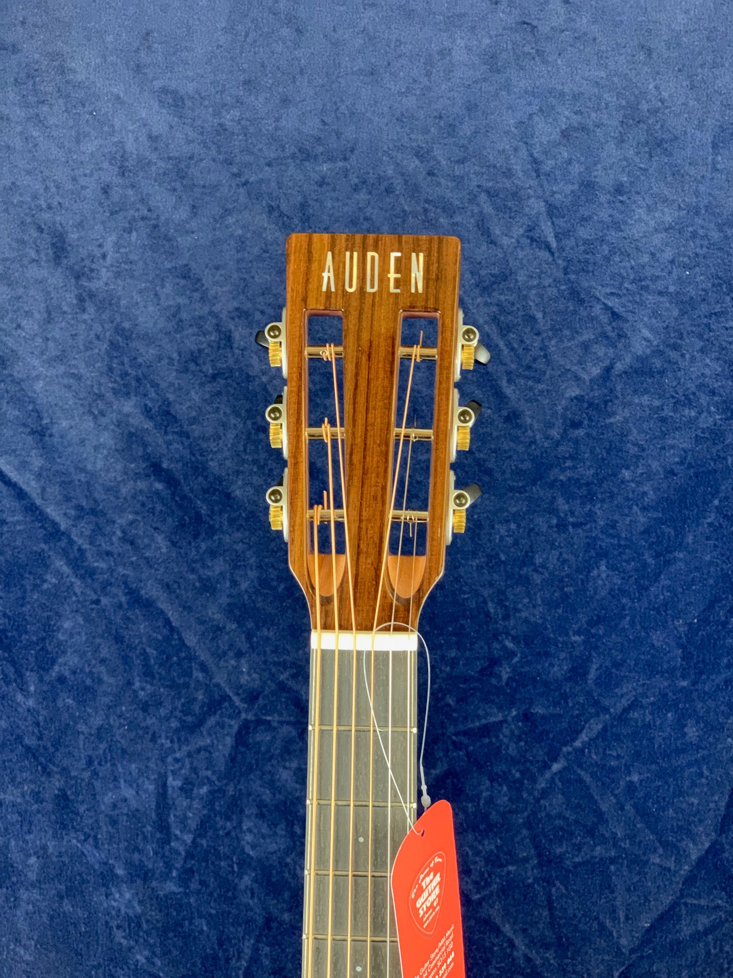 Slotted headstock of an Auden acoustic guitar with six gold tuners, wooden finish, and a red tag hanging from one string.