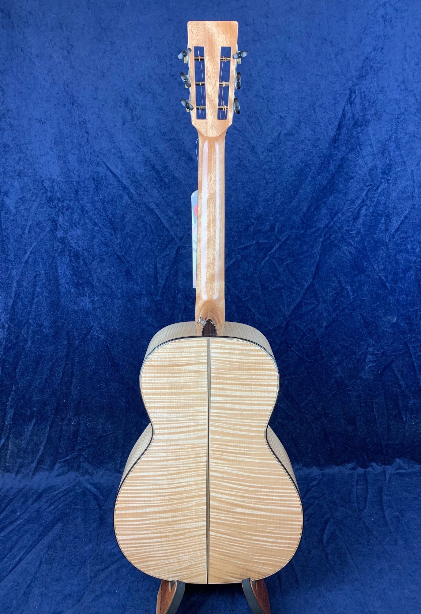 Light wood acoustic guitar shown from the back, displaying a striped grain pattern and dark trim along the edges, set against a deep blue background.