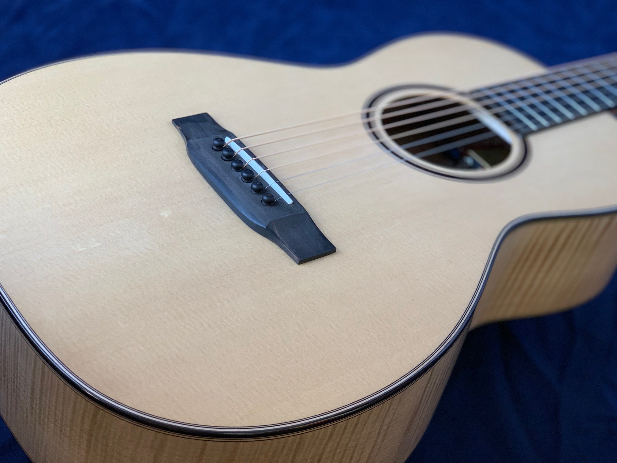 Light wood acoustic guitar with a black bridge, six strings, round sound hole, and dark binding, shown against a dark blue background.