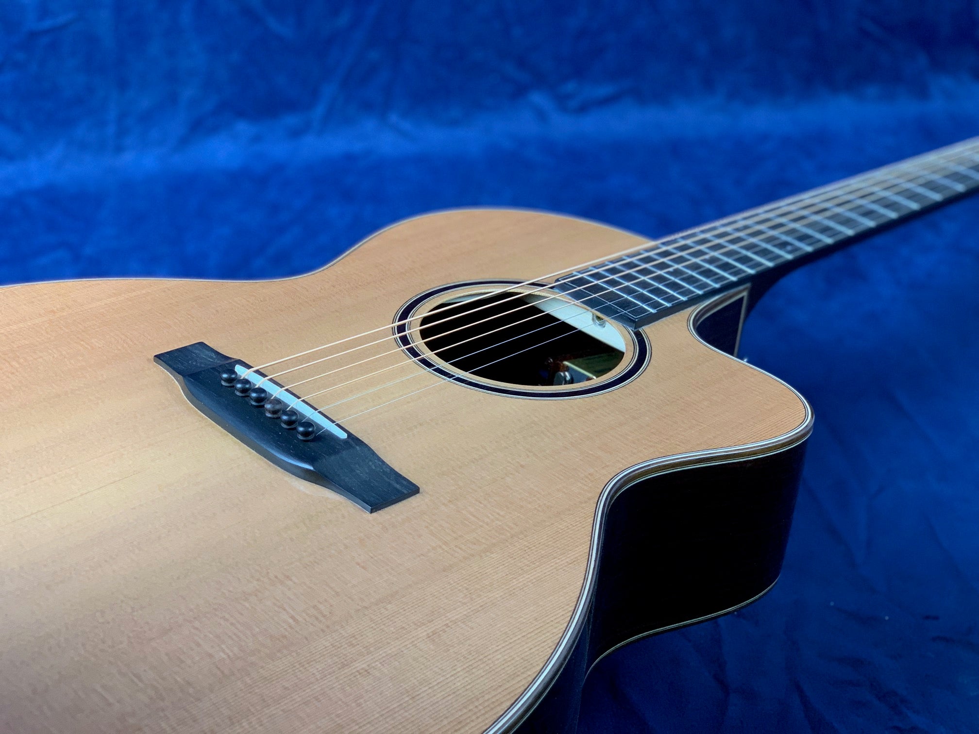 Light wood acoustic guitar with a cutaway body, six strings, and black bridge, lying on a blue textured surface.