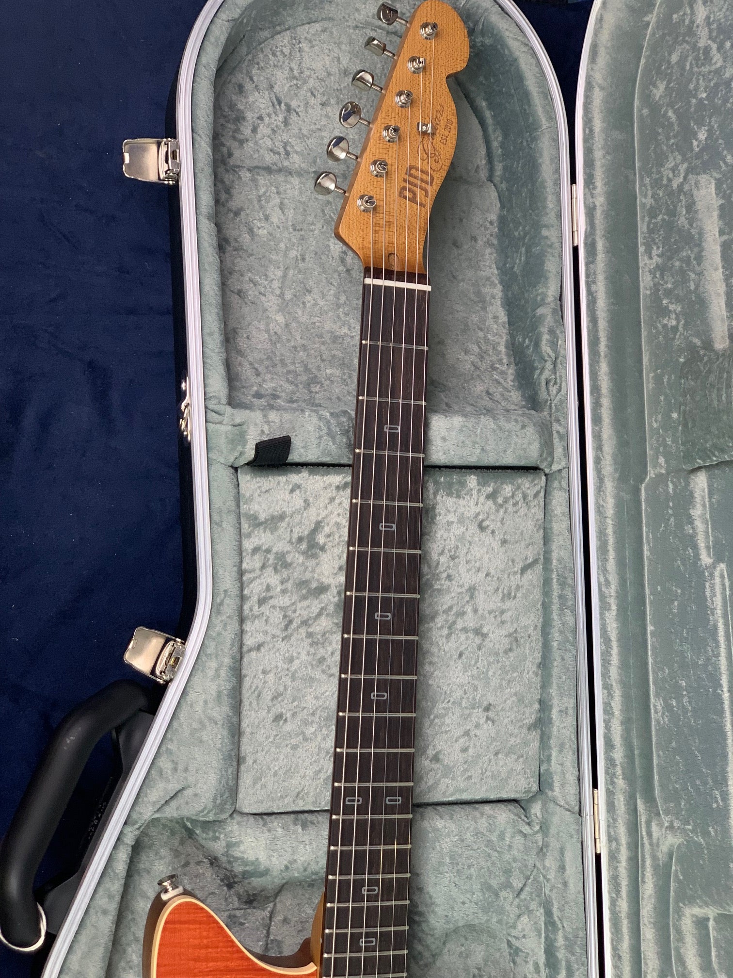 Electric guitar with a wooden neck and headstock resting in a light gray fur-lined hard case, viewed from the headstock down the neck.