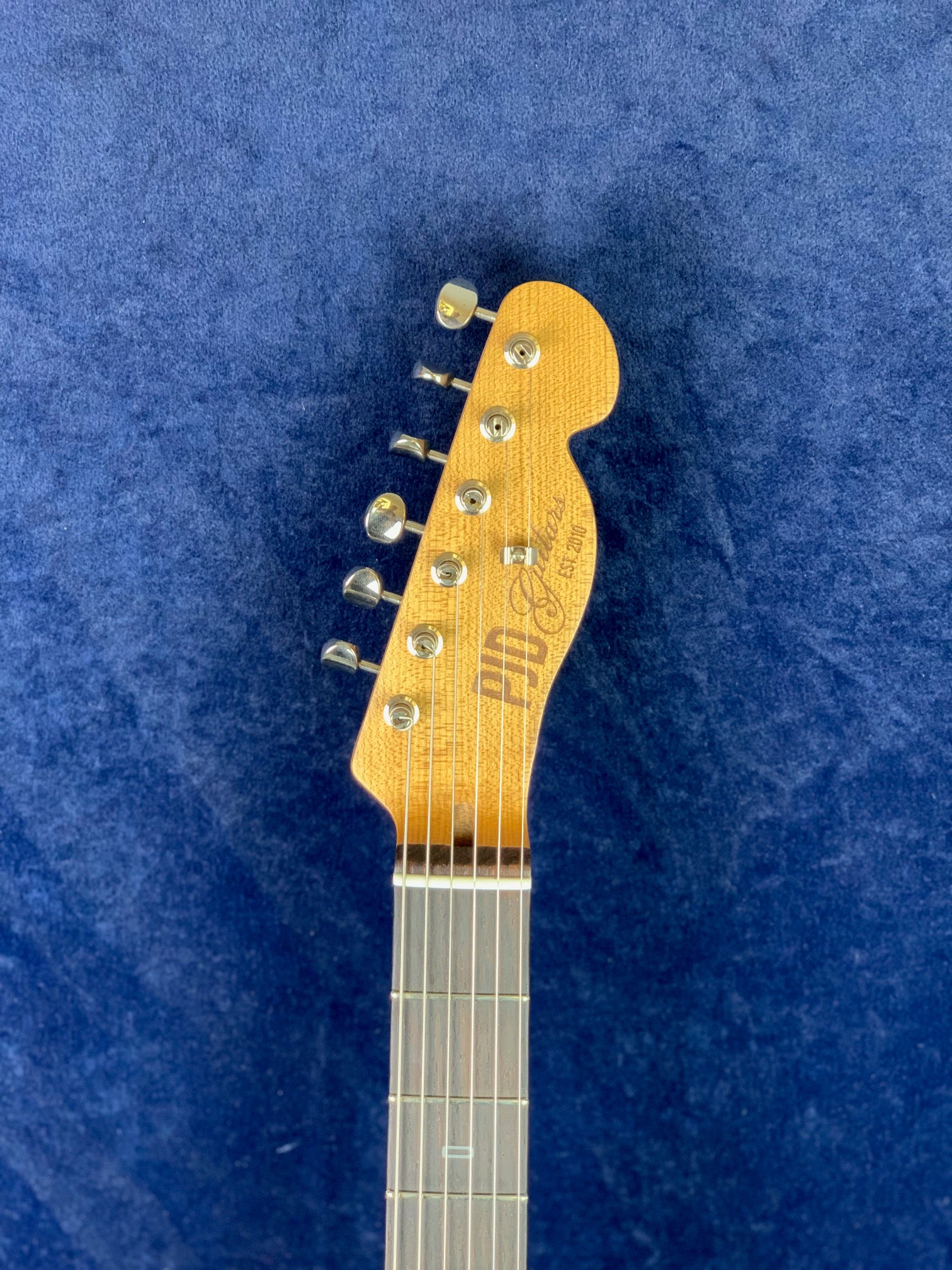 Light brown electric guitar headstock with six metal tuning pegs, brand logo, and part of a dark fingerboard against a blue background.