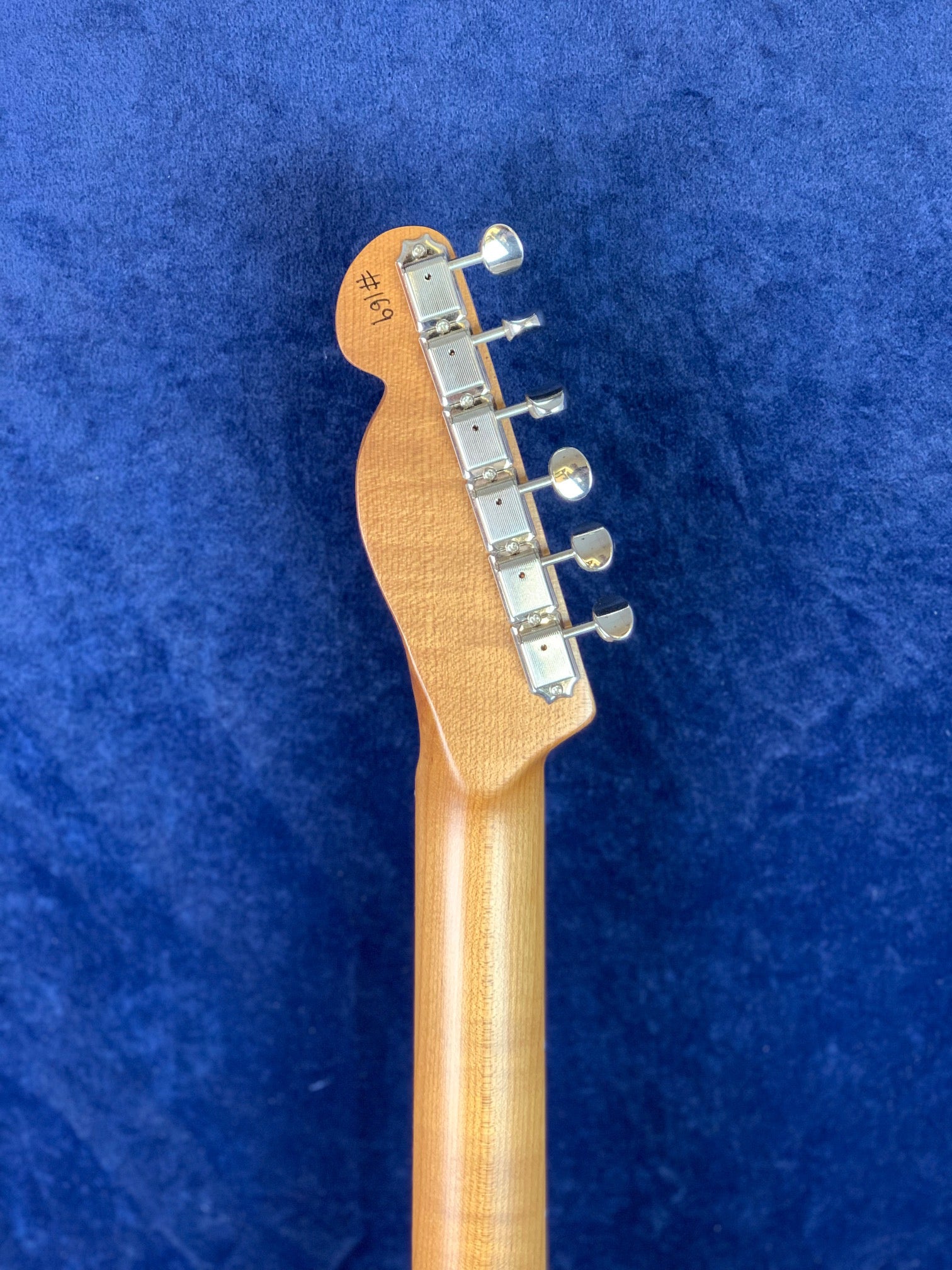 Back of an electric guitar headstock with six silver tuning pegs, light wood neck, and "#169" written on the headstock, against a blue background.